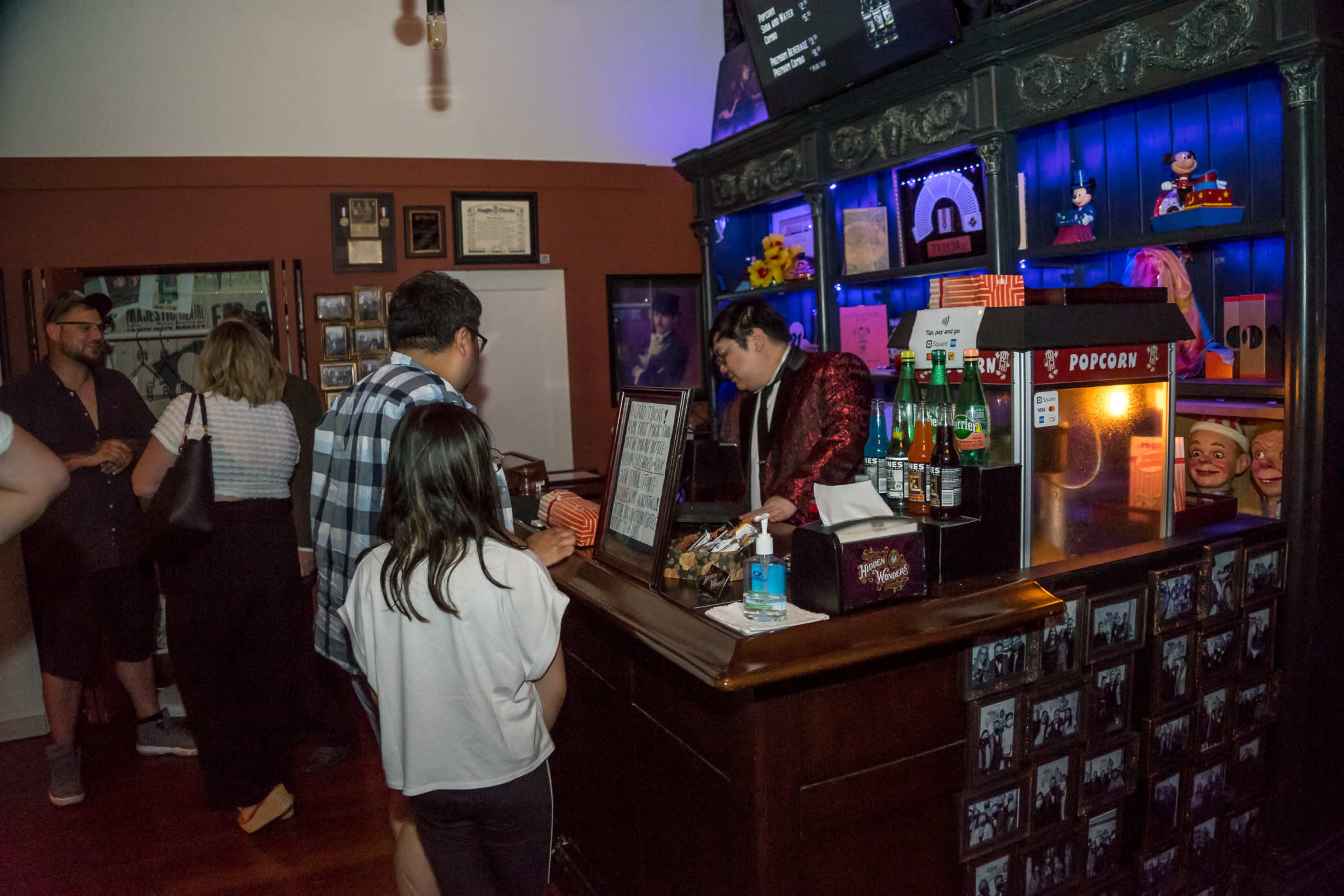 A bartender in a red jacket serves customers at a vintage-style bar illuminated by blue lighting, while a variety of memorabilia and snacks are displayed on the counter.