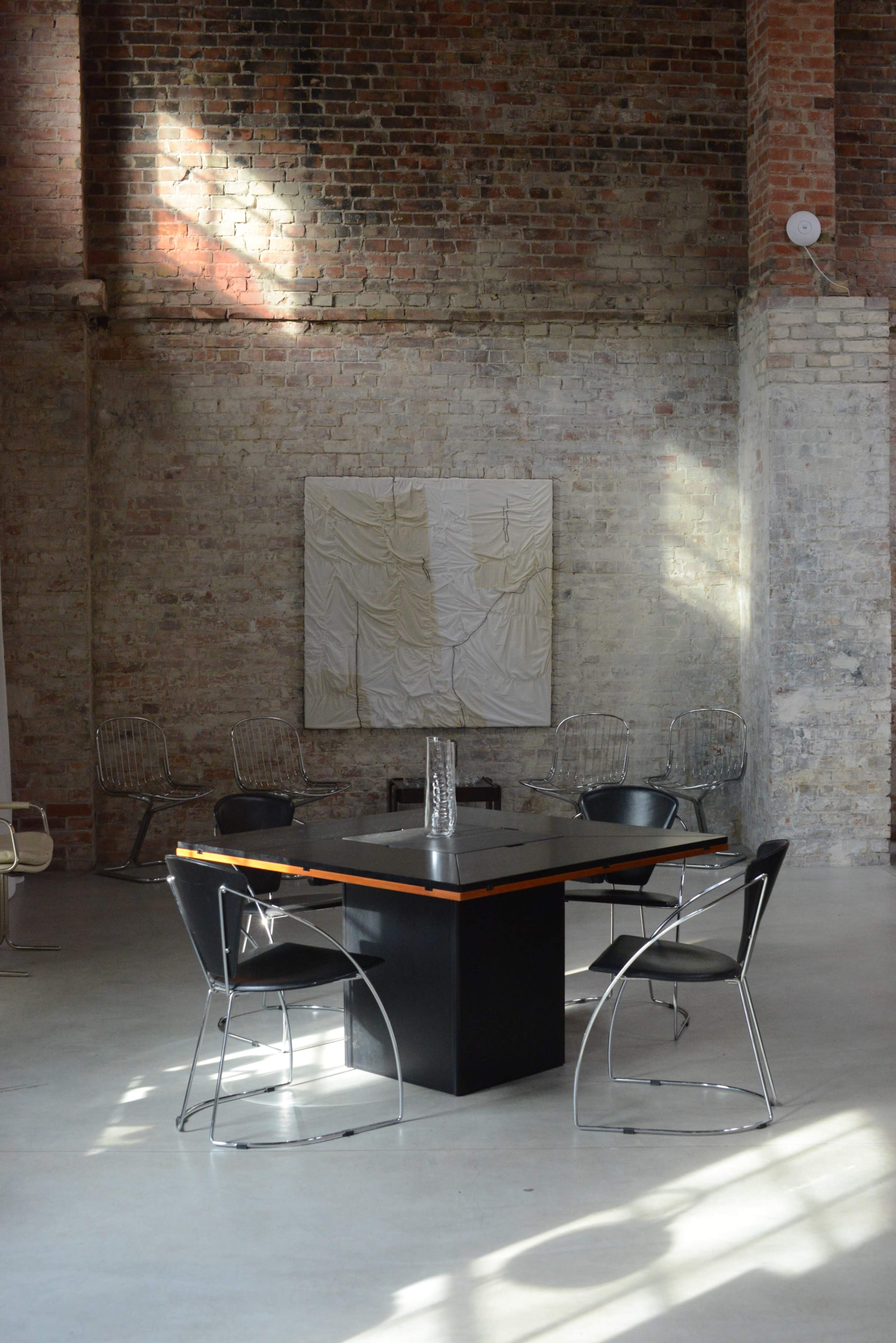 A minimalist interior features a black table surrounded by four transparent chairs, with sunlight streaming through the windows and highlighting a textured wall and artwork.