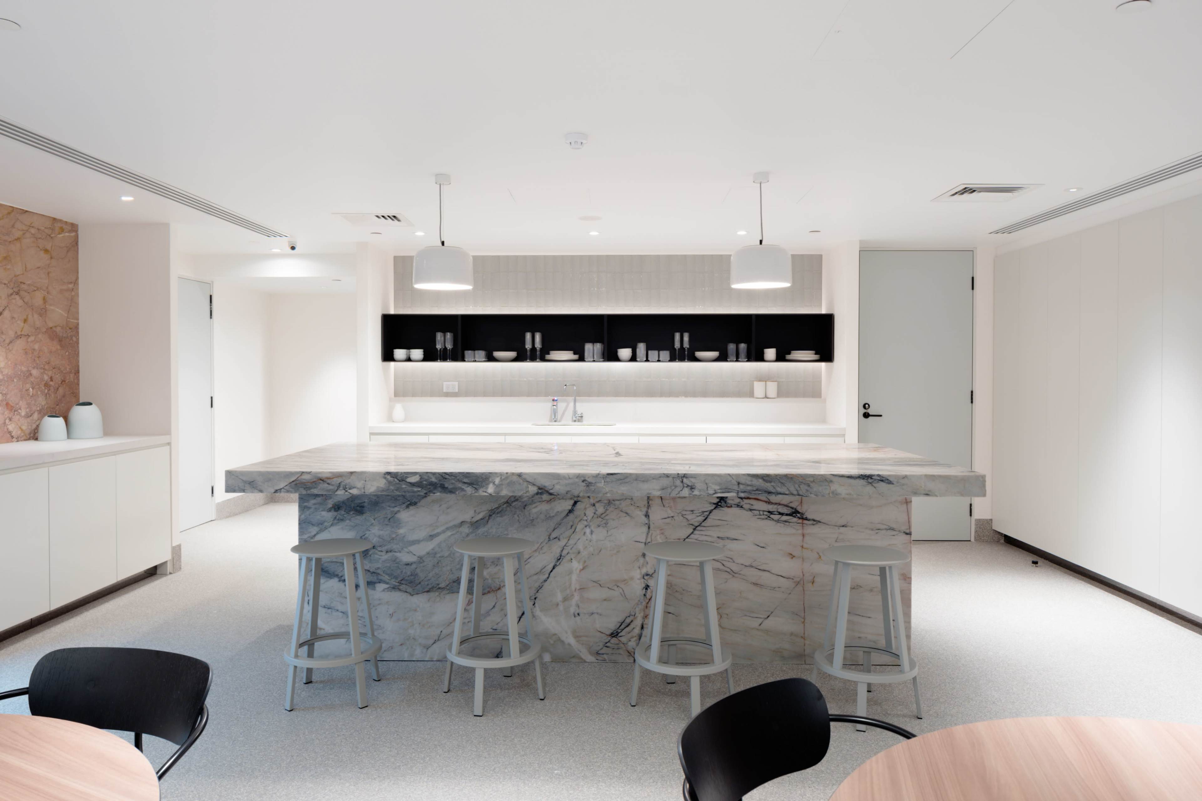 A modern kitchen with a large marble island and three stools, featuring minimalistic cabinetry and shelves with small containers.