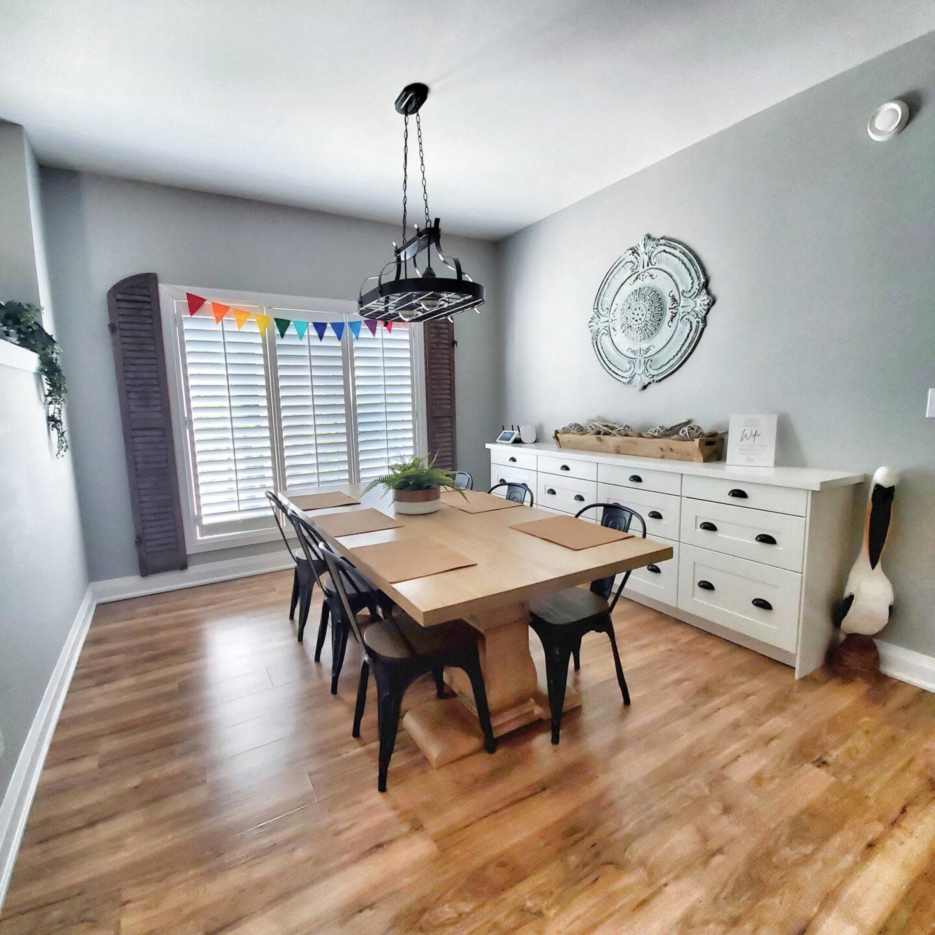 A dining room features a large wooden table surrounded by black chairs, with a sideboard and window adorned with decorative banners.