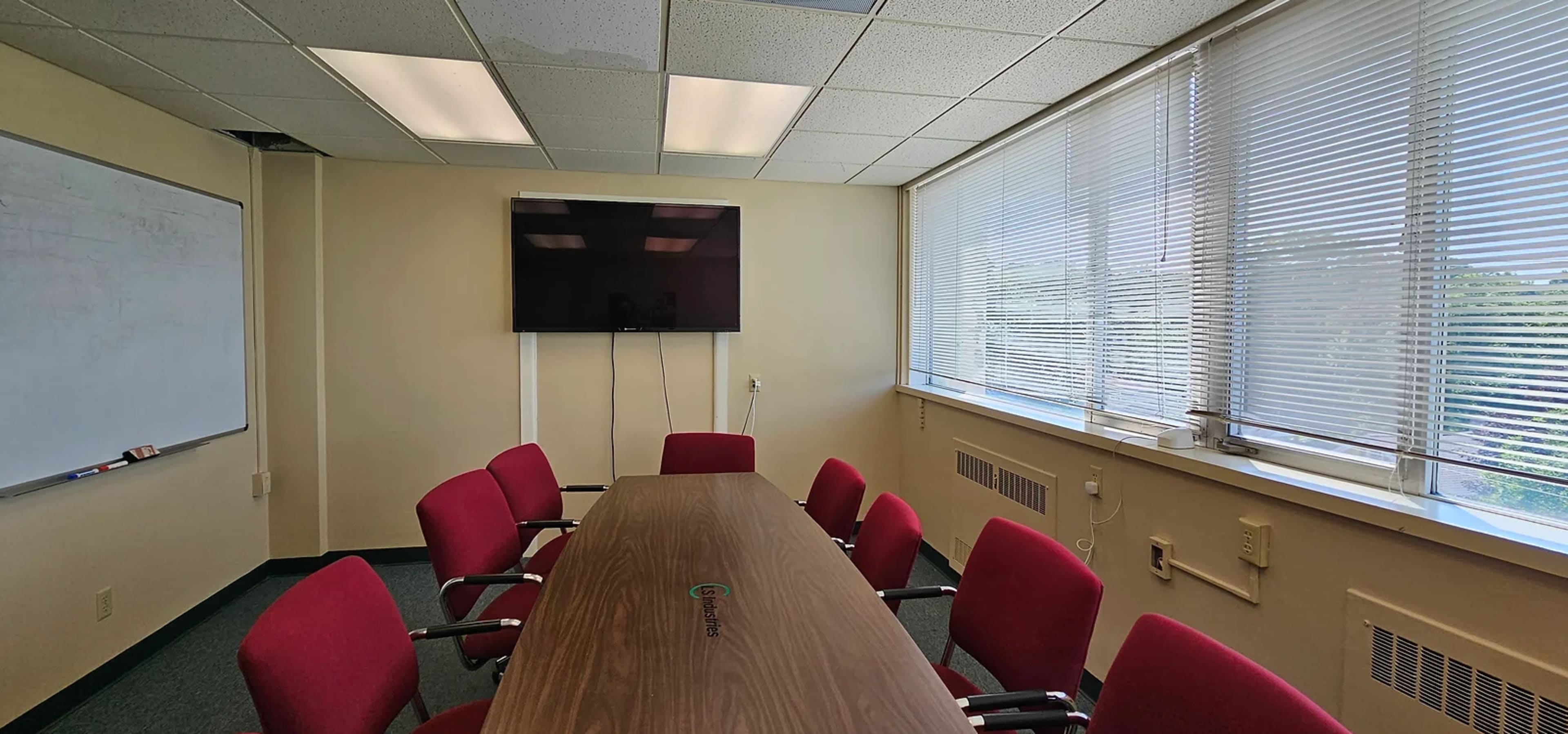 A conference room features a long wooden table surrounded by red chairs, with a wall-mounted TV and large windows covered by blinds.