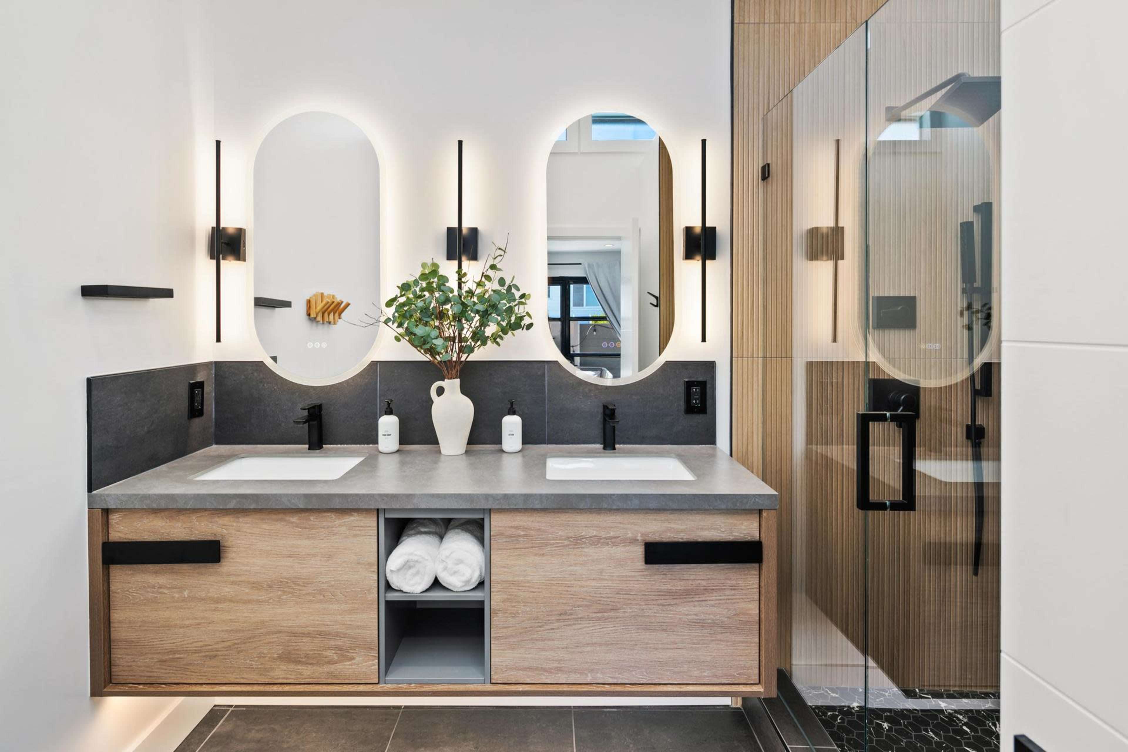 The image shows a modern bathroom featuring a dual-sink vanity with a concrete countertop, wooden cabinets, and two round mirrors illuminated by wall sconces, alongside a glass shower enclosure.