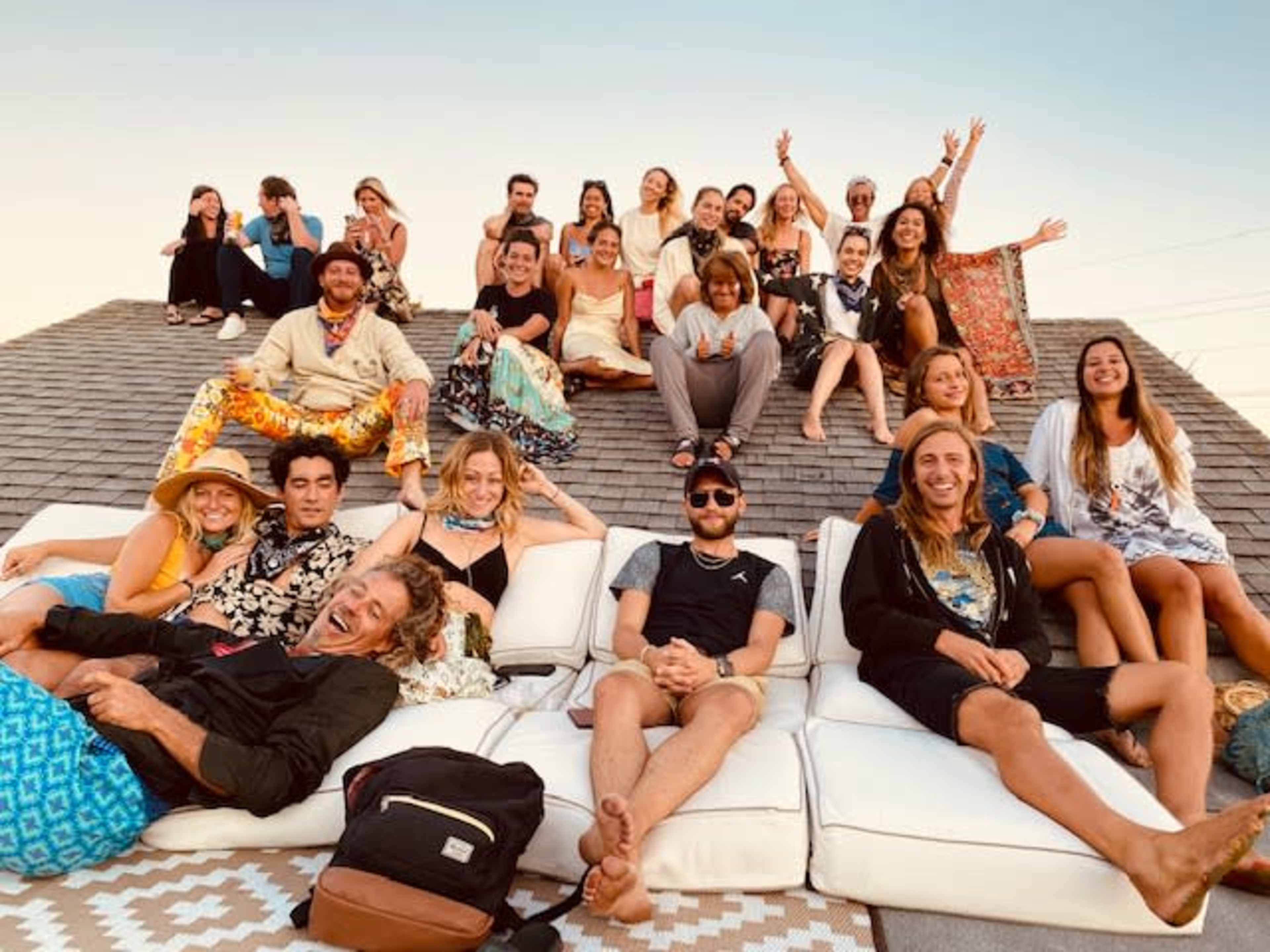 A diverse group of people gathers on a rooftop, lounging on large cushions and enjoying the sunset.