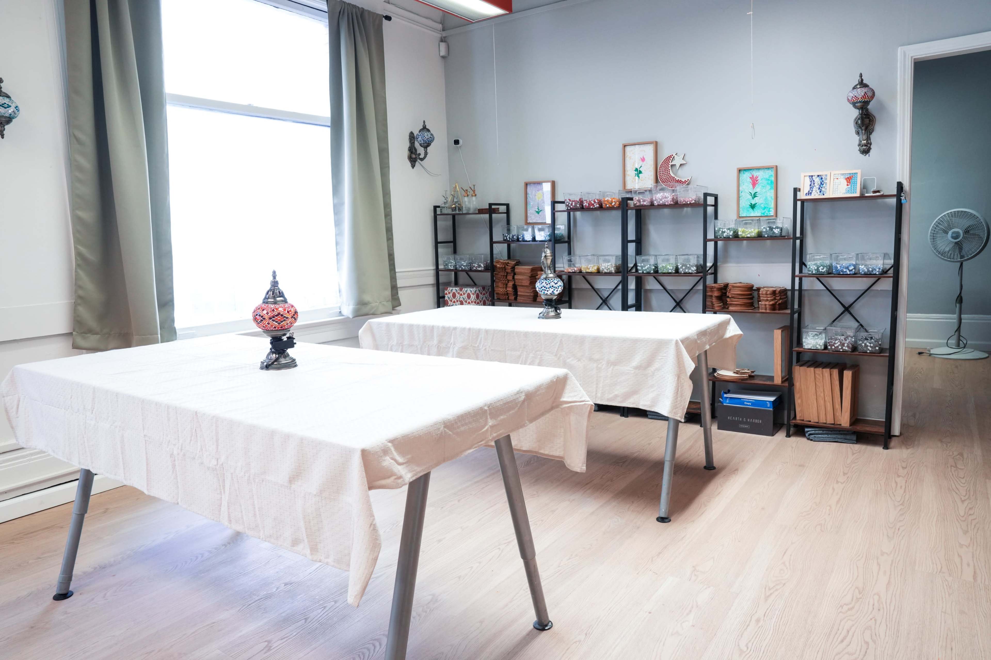 The image shows a bright, spacious room with two tables covered in white cloths, decorated with ornate lamps and shelves filled with various items in the background.