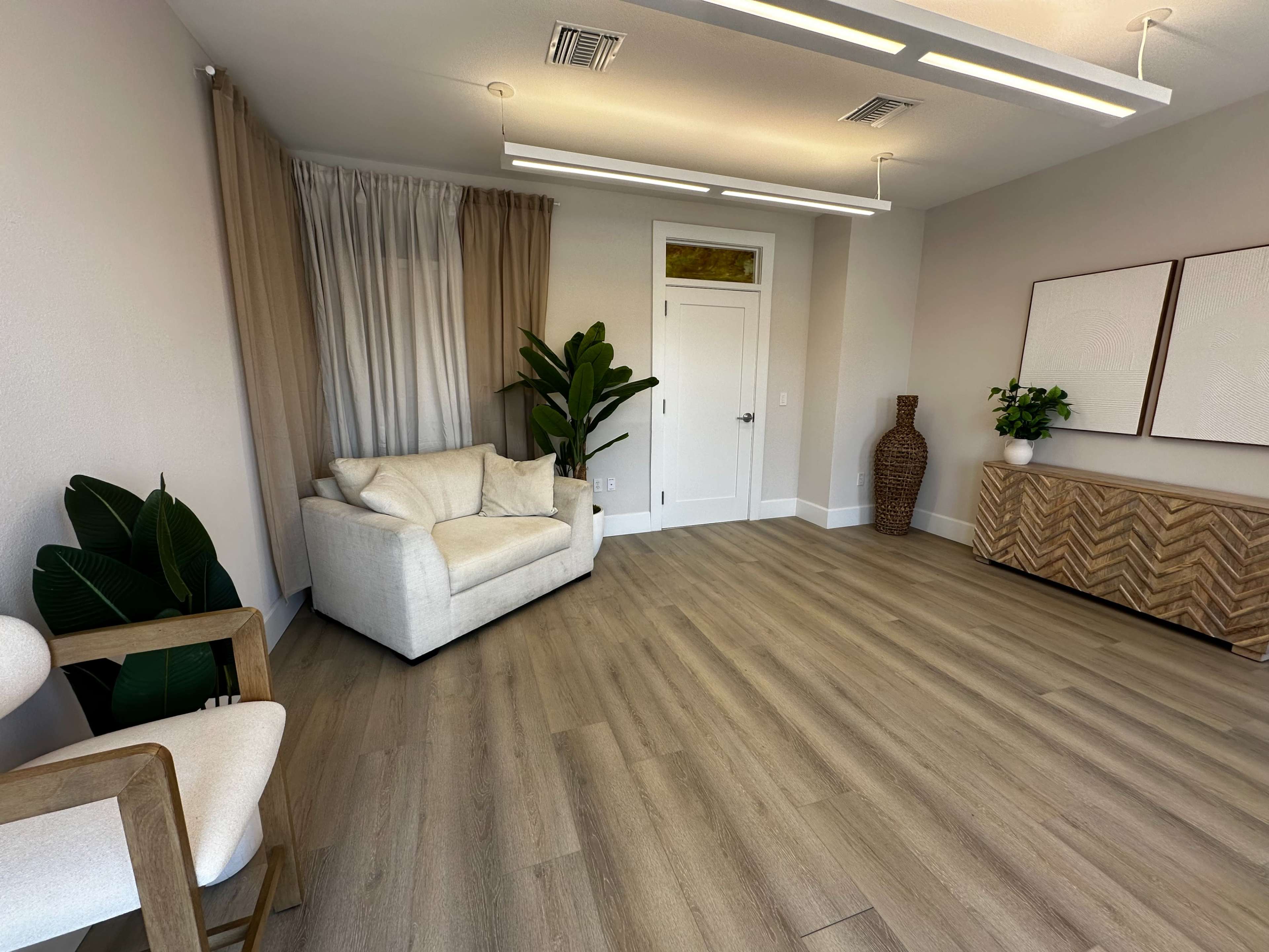 A spacious, modern room featuring a white armchair, a light-colored sofa, a tall plant, a decorative vase, and two framed artworks on a wooden sideboard.