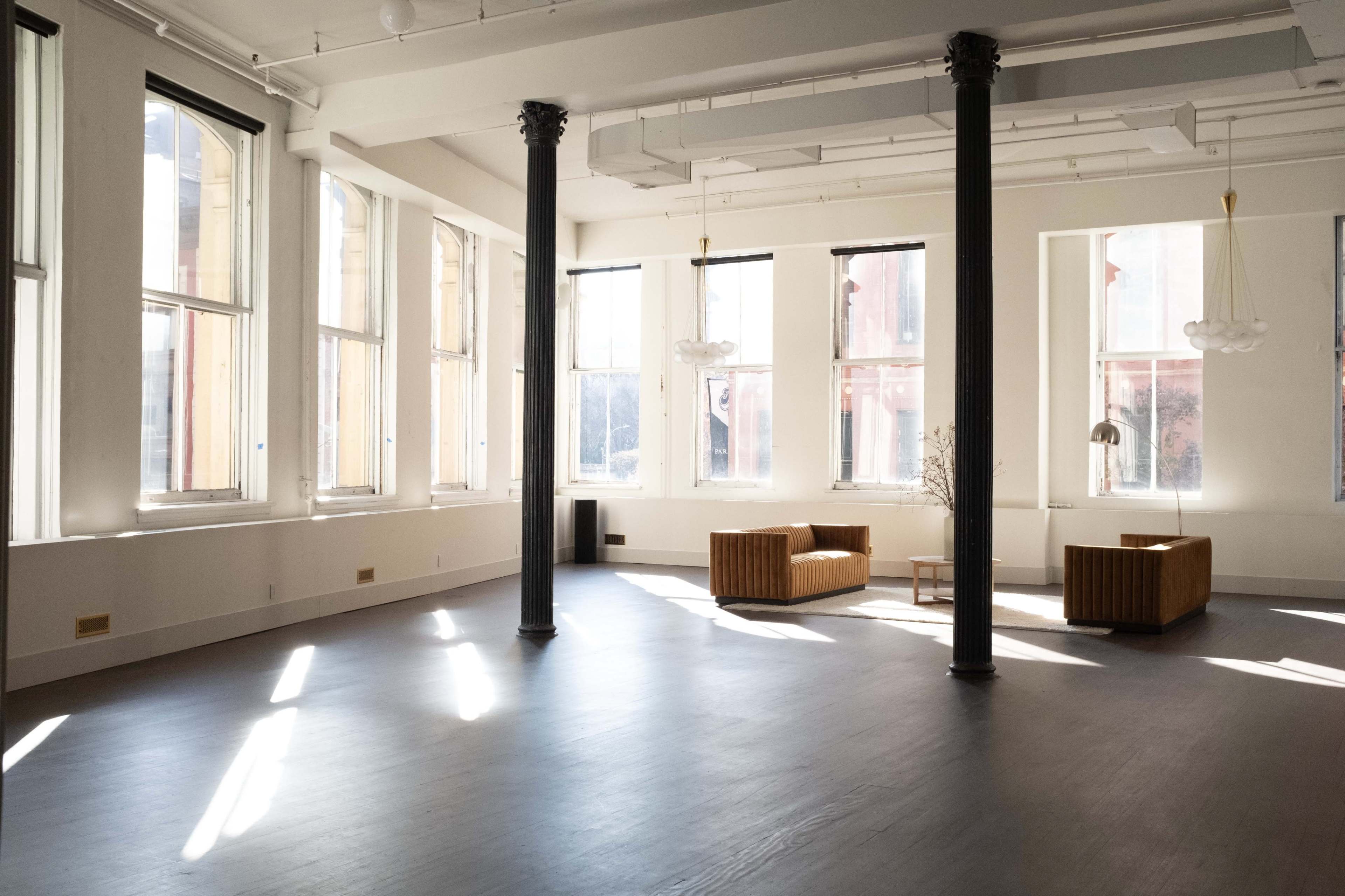 The image shows a spacious, sunlit interior with large windows, two brown seating areas, and high ceilings supported by black columns.
