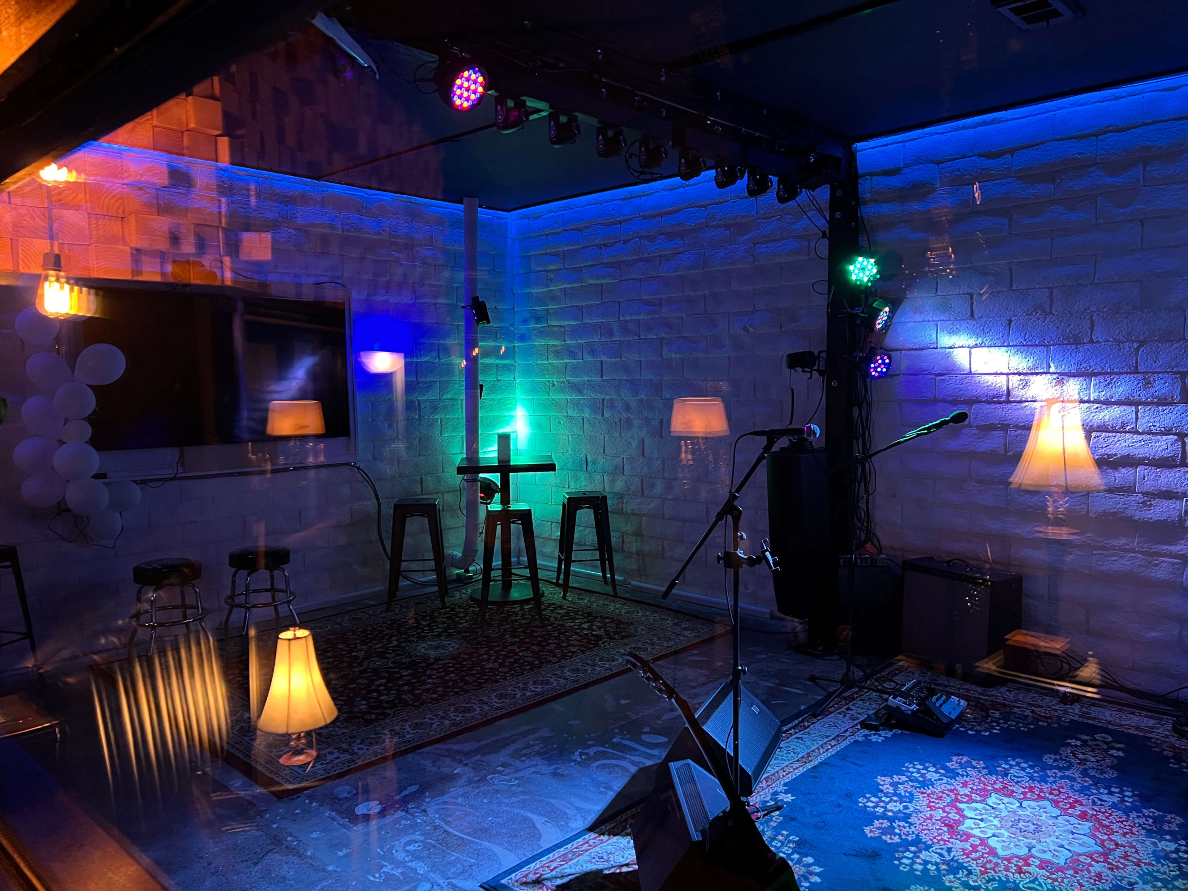 A small performance space with colorful lighting, several stools, and musical equipment, featuring a patterned rug and floor lamps.