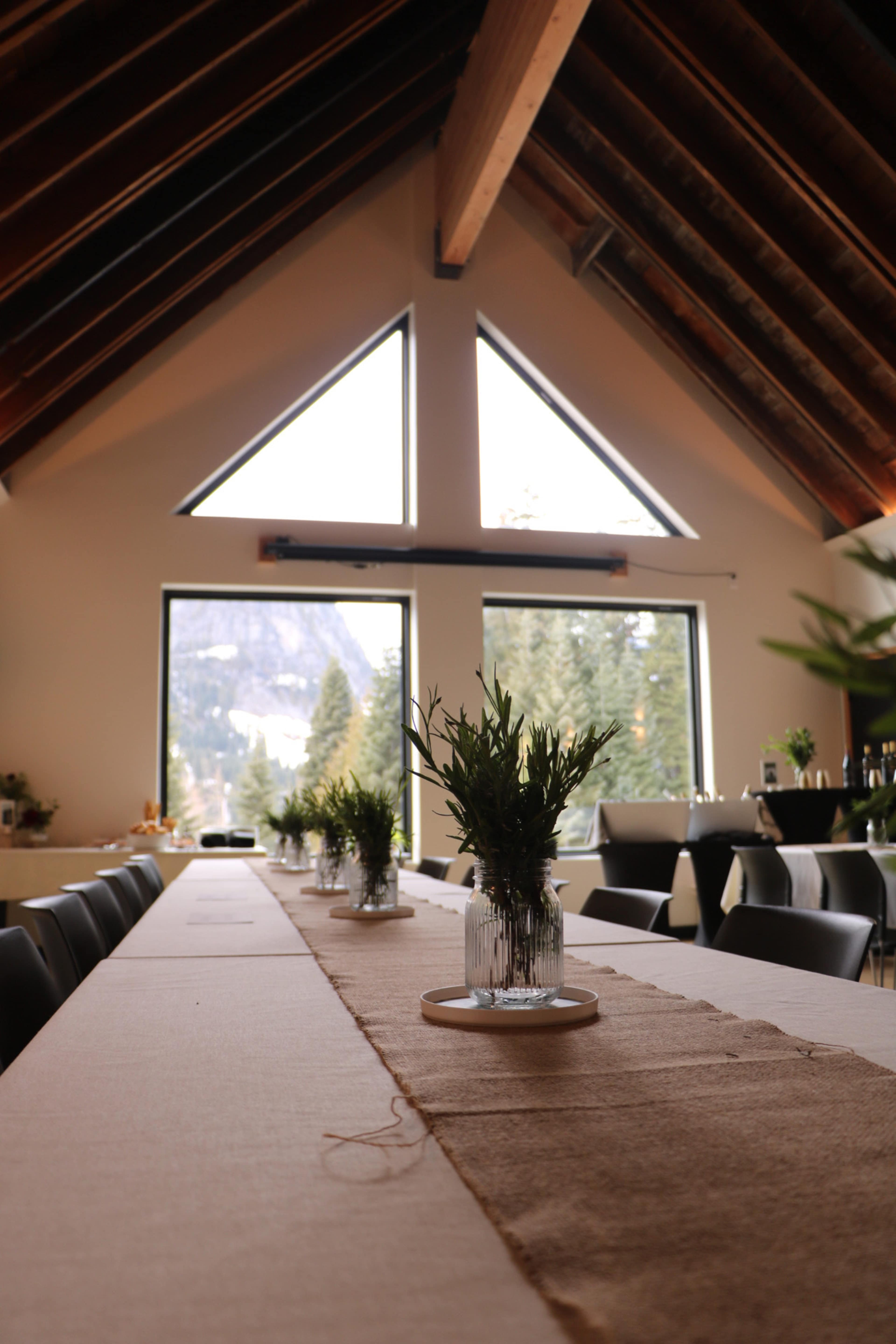 A long dining table is set in a room with a vaulted ceiling and large windows overlooking a mountainous landscape.