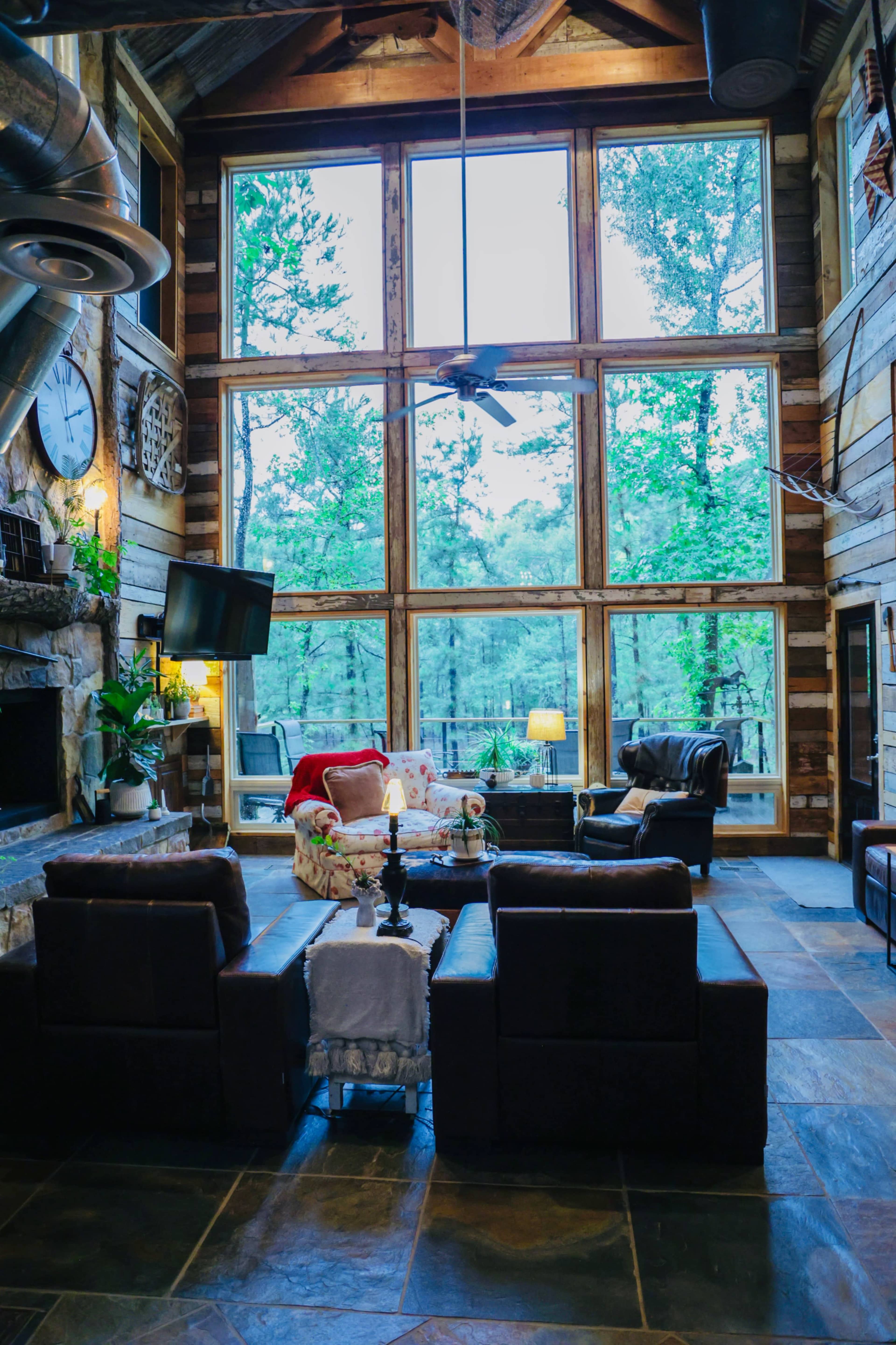 A spacious living room with large windows overlooking a forested area, featuring a stone fireplace and comfortable seating arrangements.