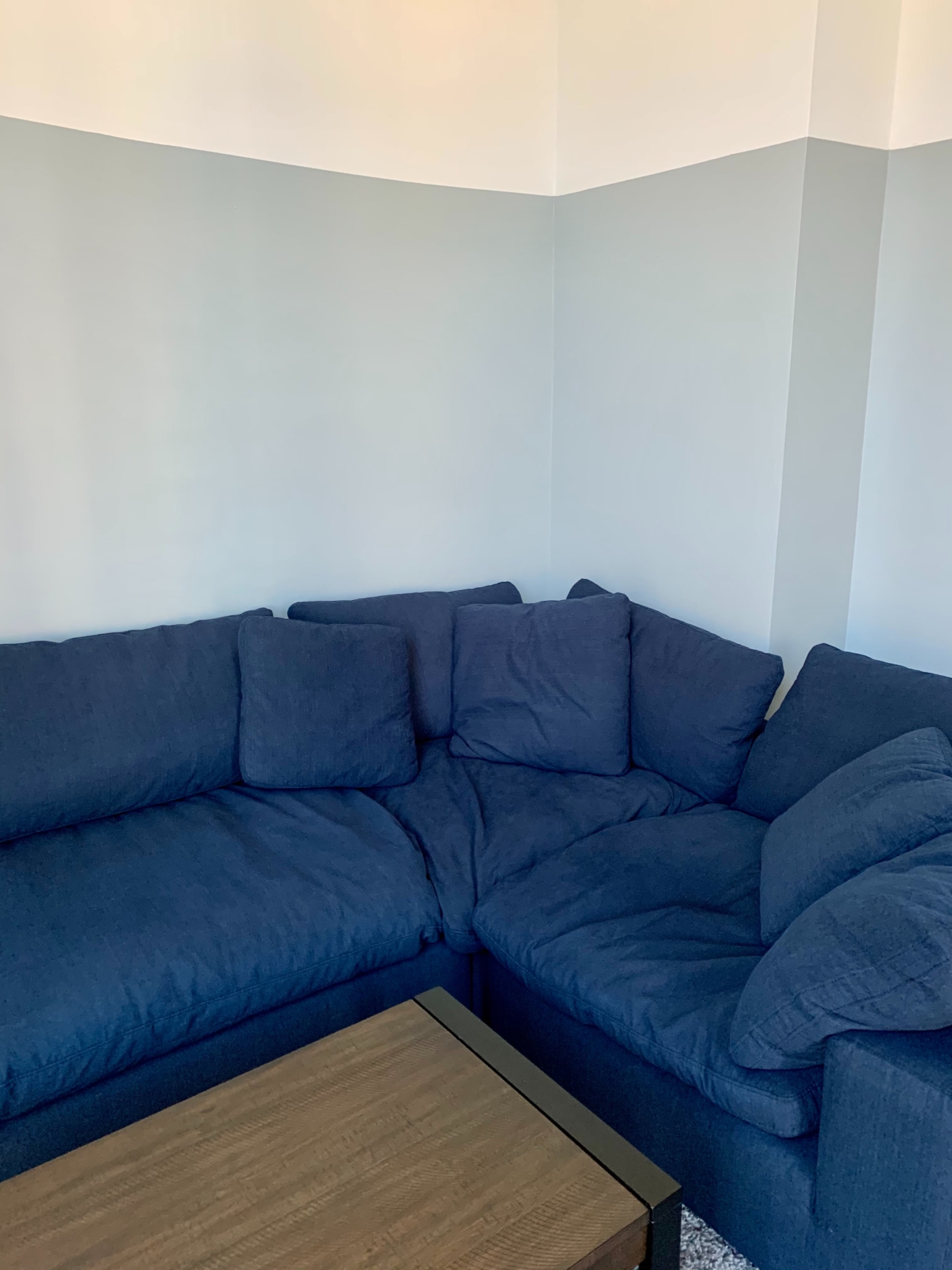 The image shows a corner sectional sofa covered in dark blue fabric, positioned near a light-colored wall, accompanied by a wooden coffee table.
