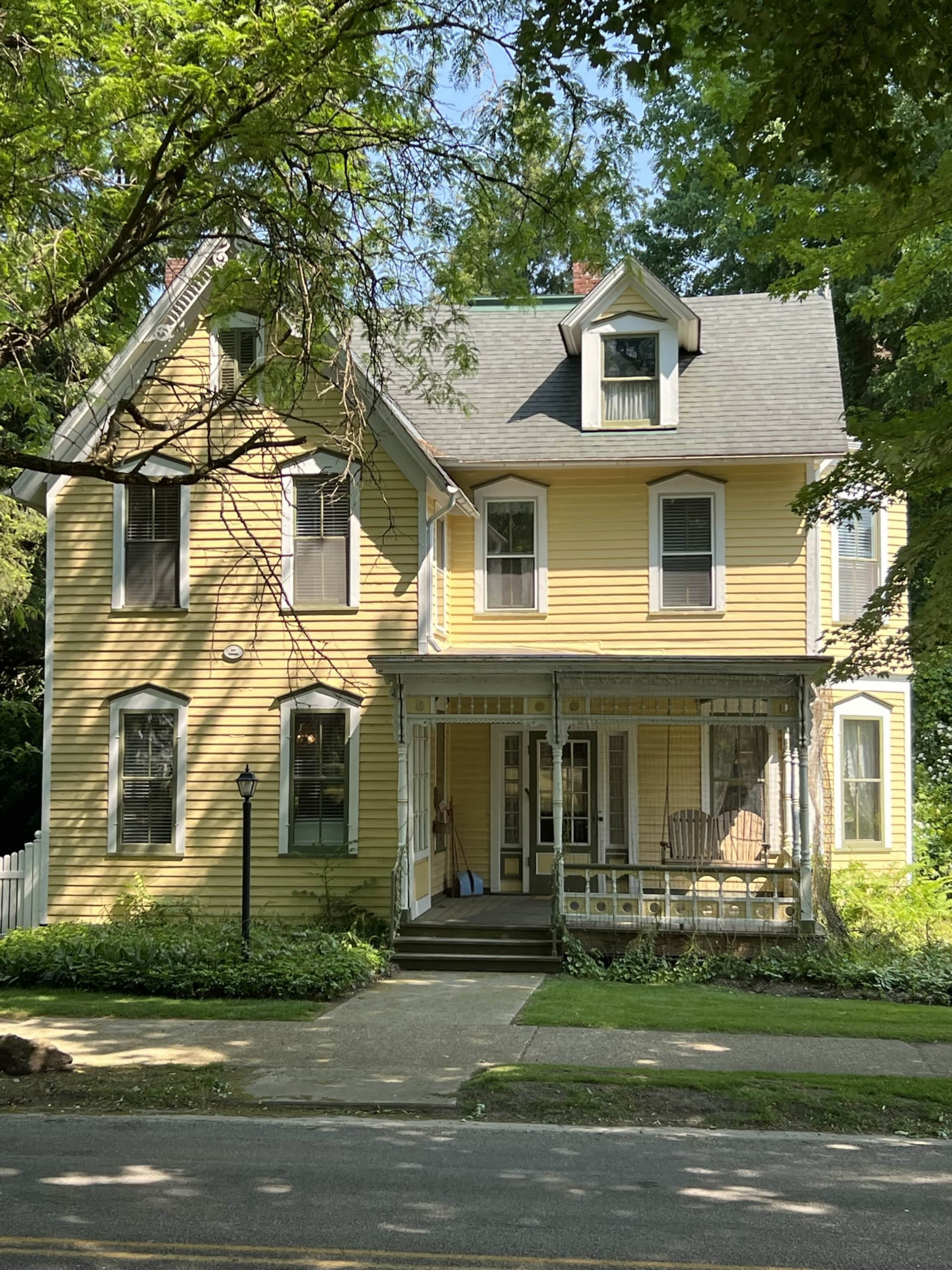 A yellow Victorian-style house with a front porch and multiple gabled roofs sits among lush green trees.
