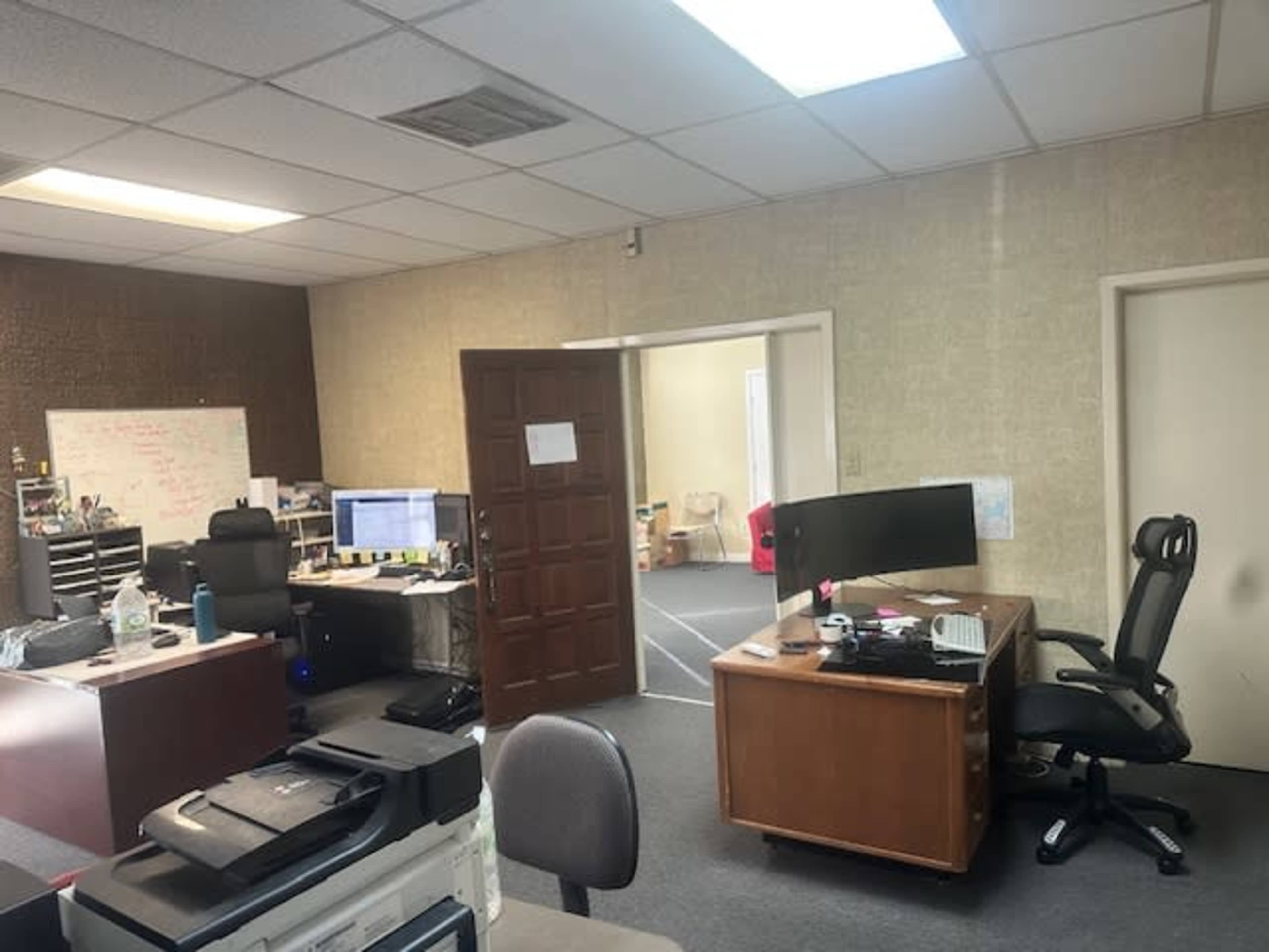 The image shows a spacious office with two workstations, a large desk, and a doorway leading to another room.