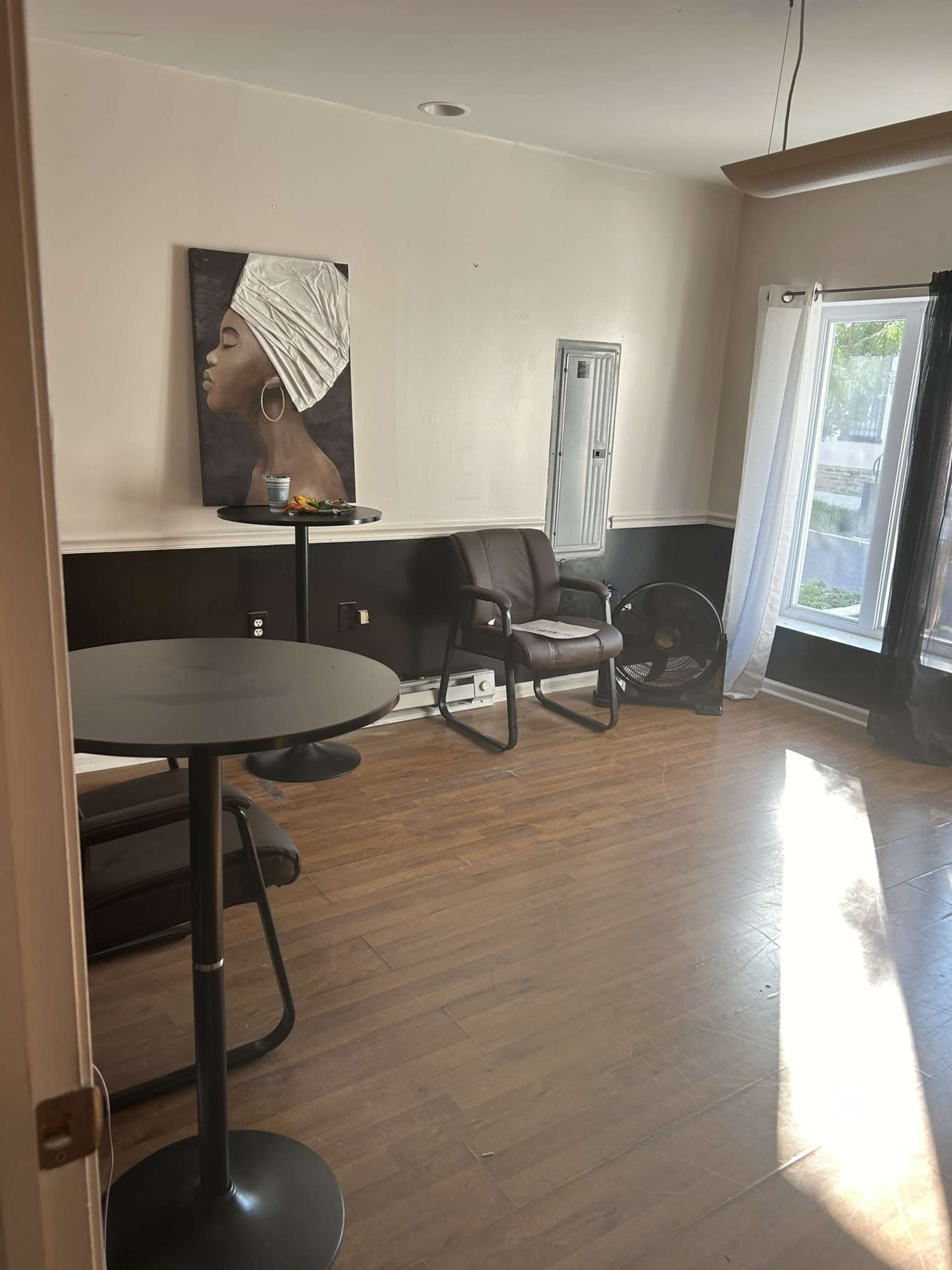 The image shows a well-lit room with a dark wooden floor, featuring a table and two brown chairs, a fan, and a wall art piece of a woman with a head wrap.