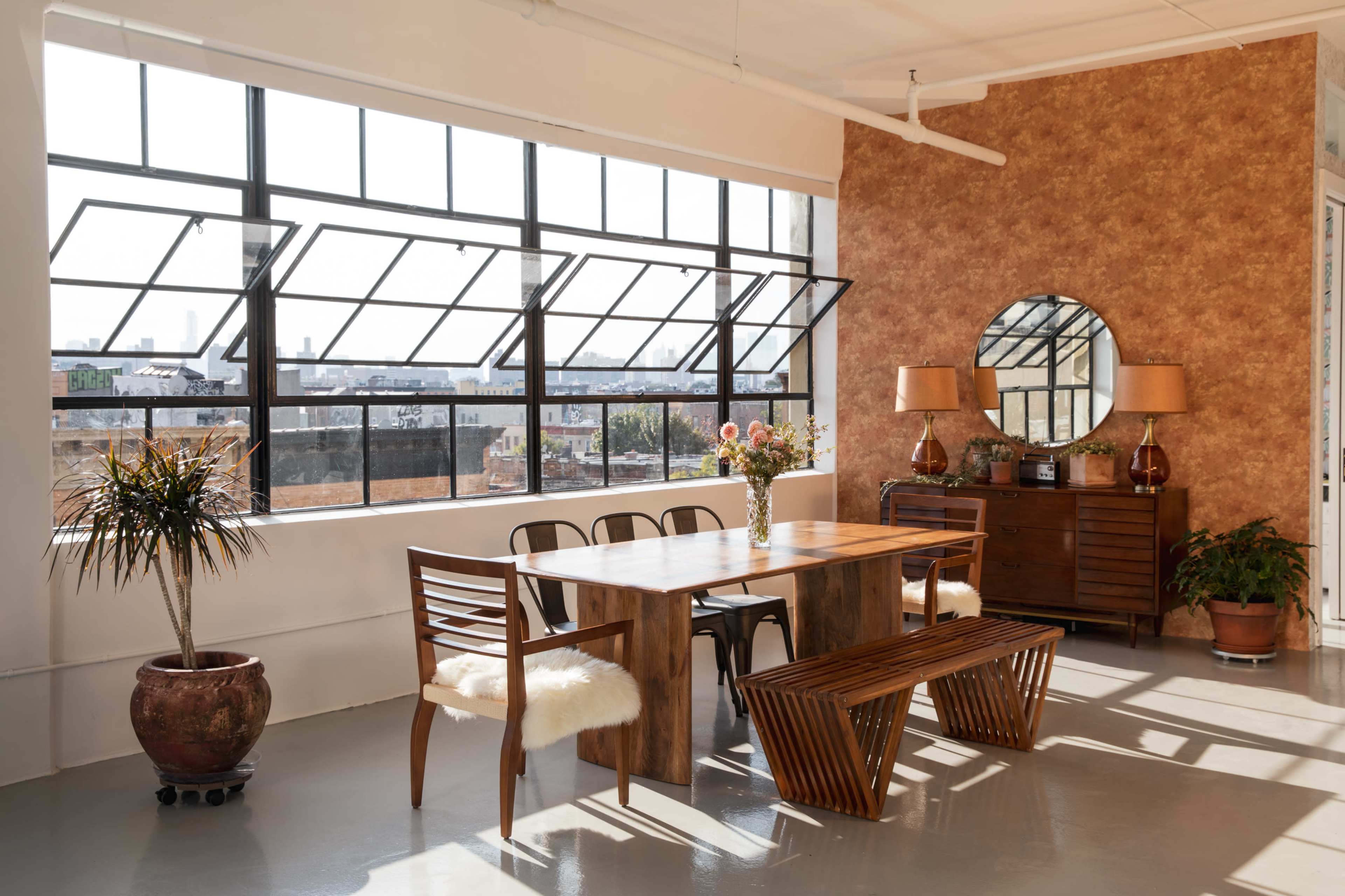 A spacious dining area with a wooden table, black chairs, a bench, and large windows letting in natural light, surrounded by plants and decorative furnishings.