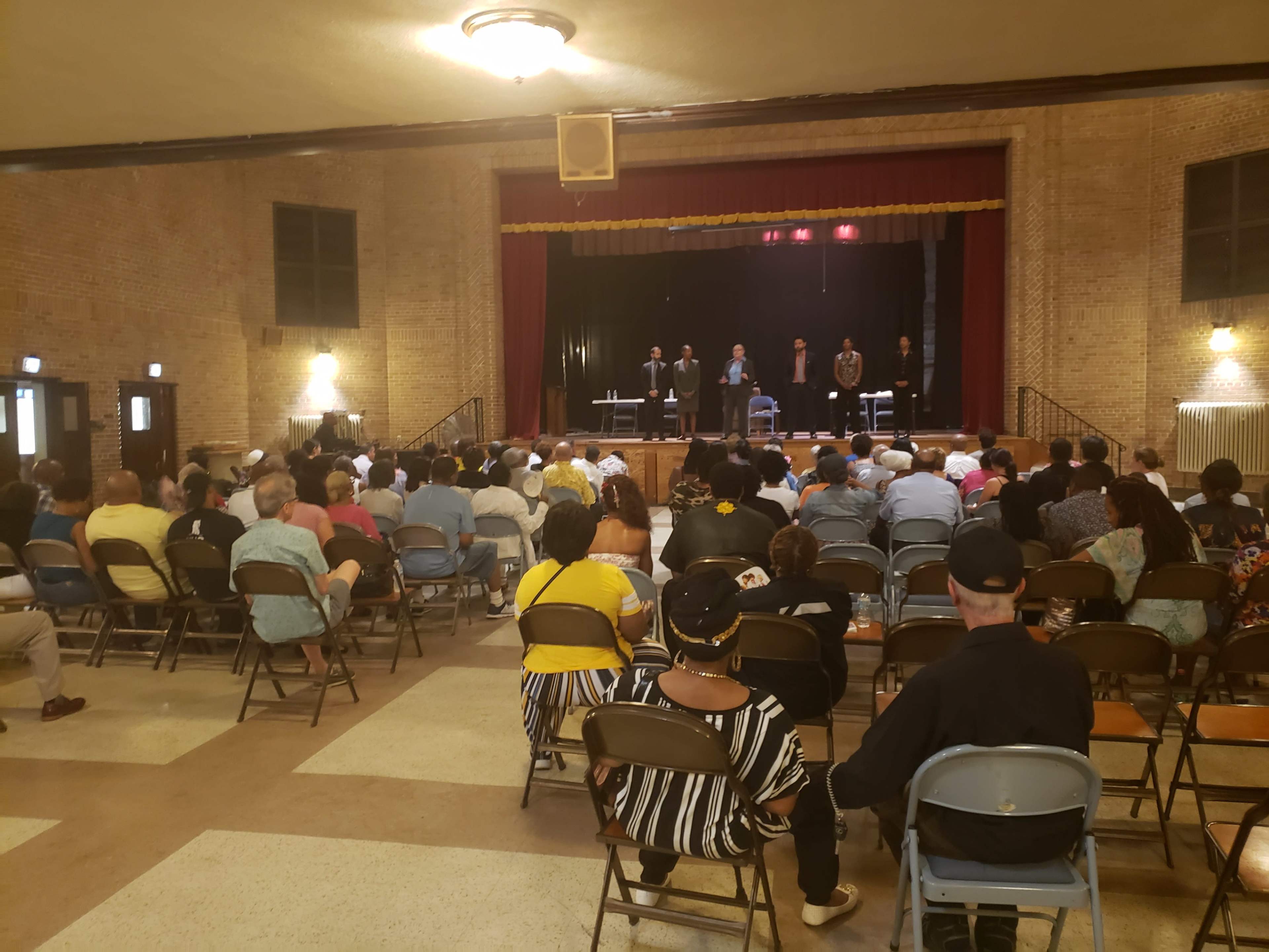 A large audience is seated in a hall, facing a stage where several speakers stand behind tables.