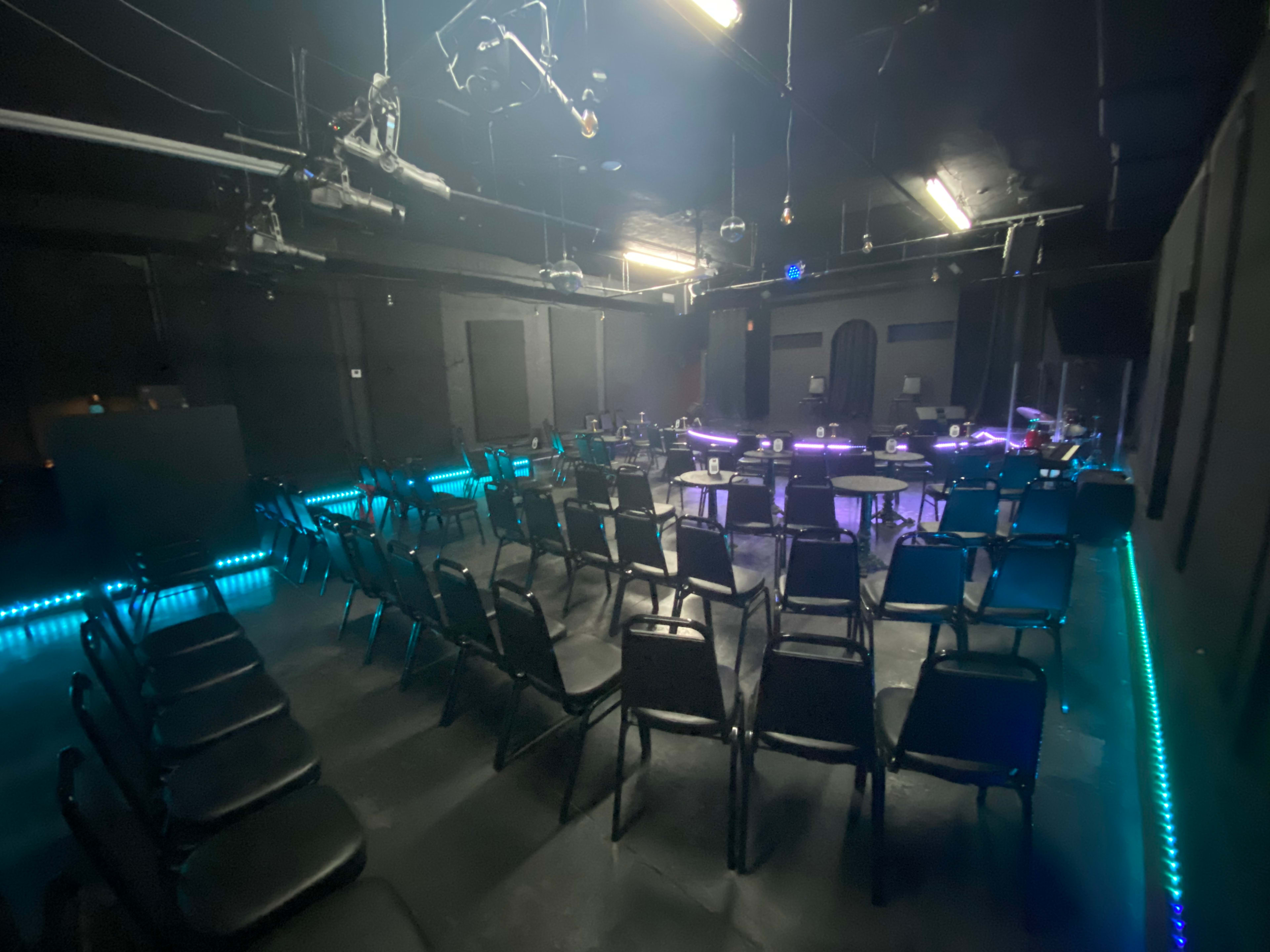 A dimly lit performance space with rows of black chairs arranged in front of a small stage adorned with colorful lights.