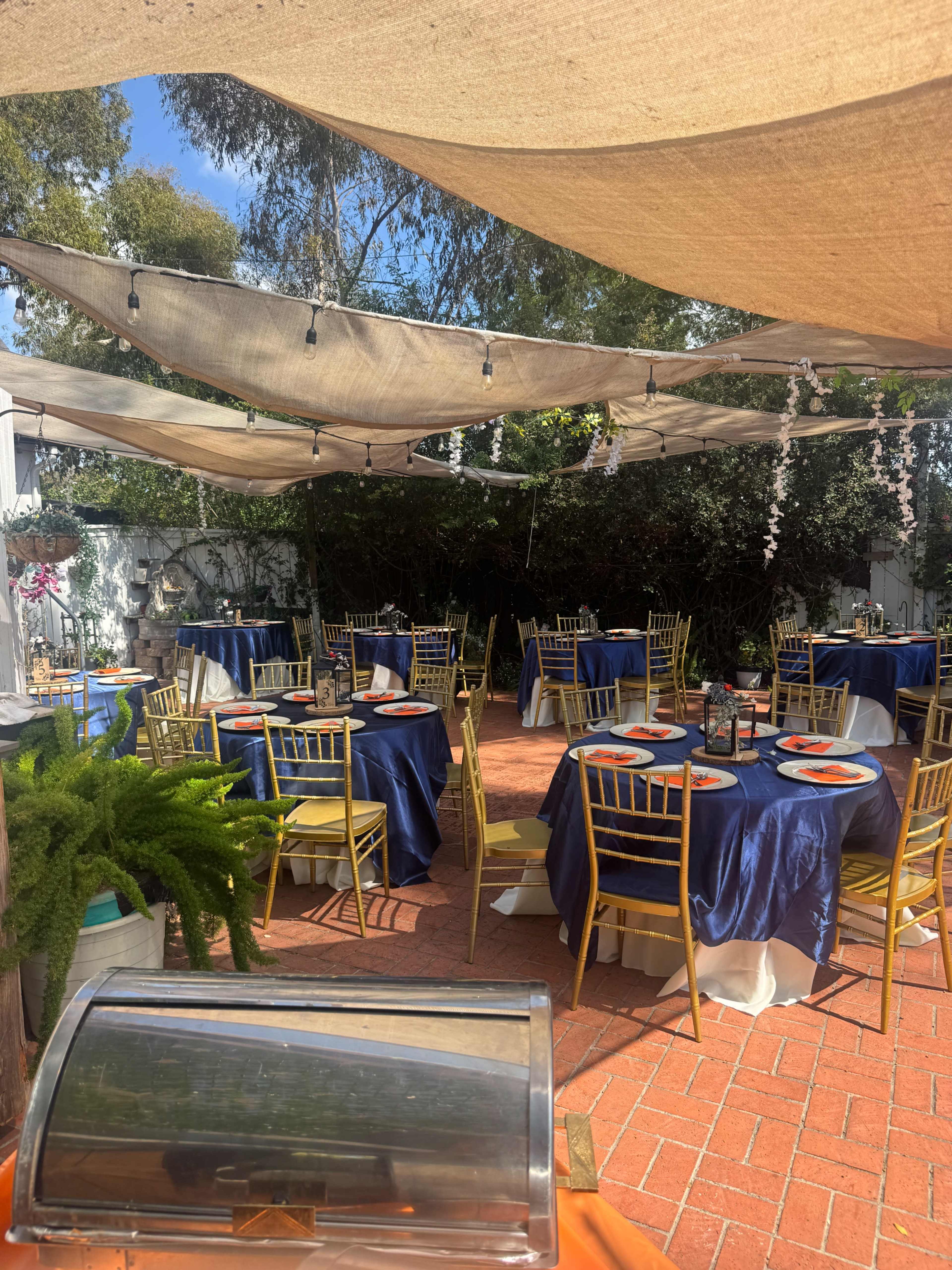 The image shows an outdoor dining area set with round tables covered in navy blue tablecloths, each adorned with decorative plates and surrounded by gold chairs, under a canopy of fabric and string lights.