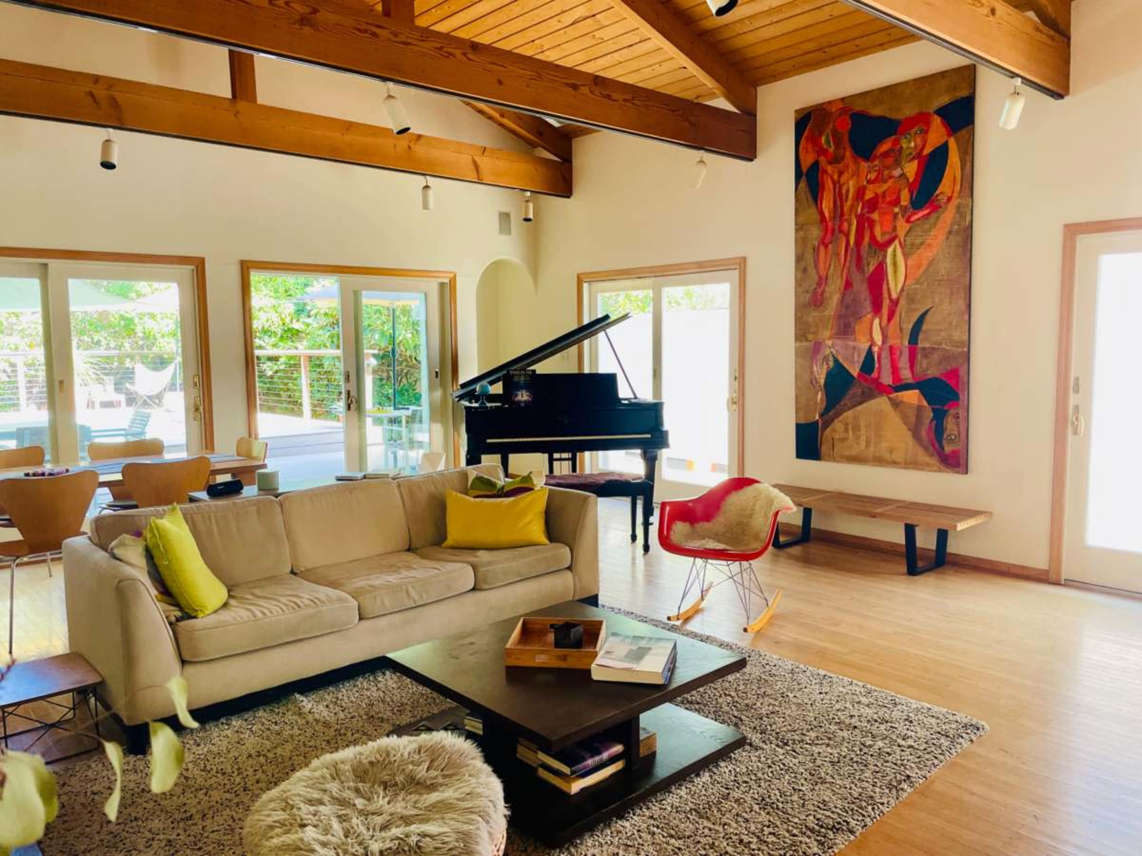 A modern living room features a beige sofa, a grand piano, a coffee table, and a large abstract painting on the wall, with glass doors leading to an outdoor area.