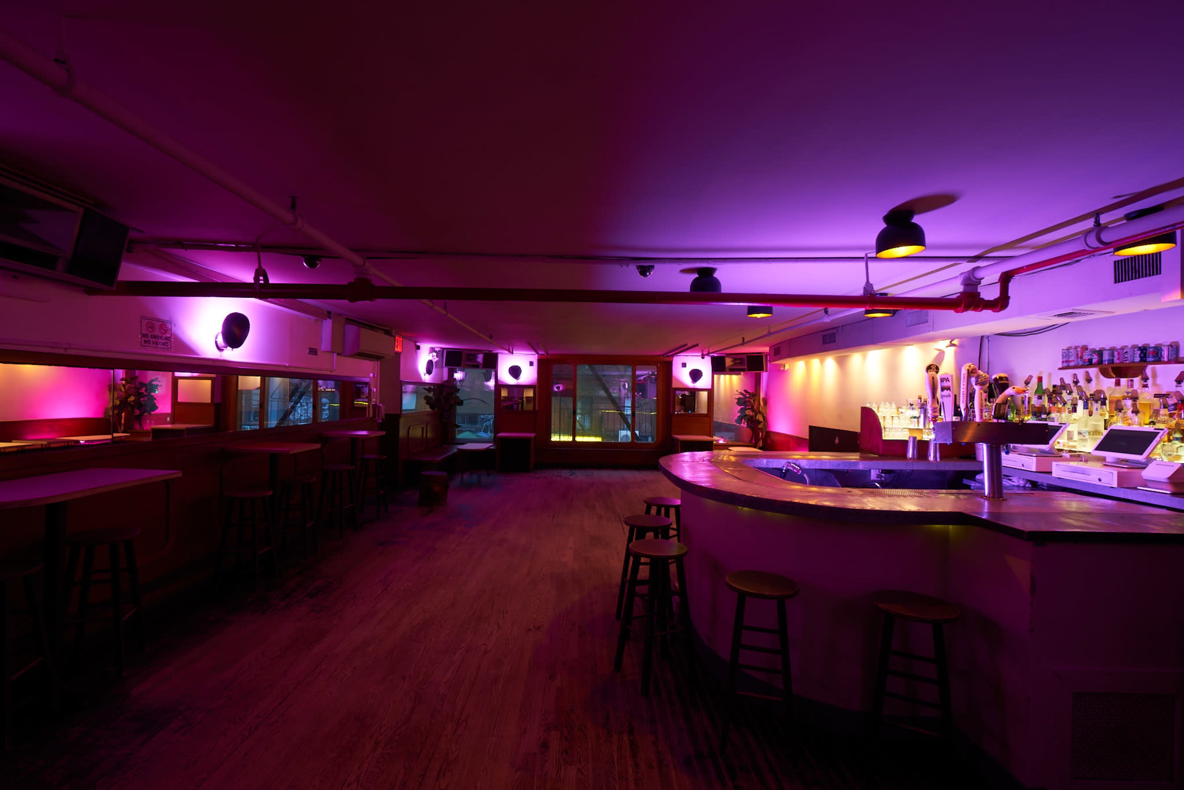 The image shows an empty bar with purple lighting, wooden flooring, and a long bar counter illuminated by overhead lights.