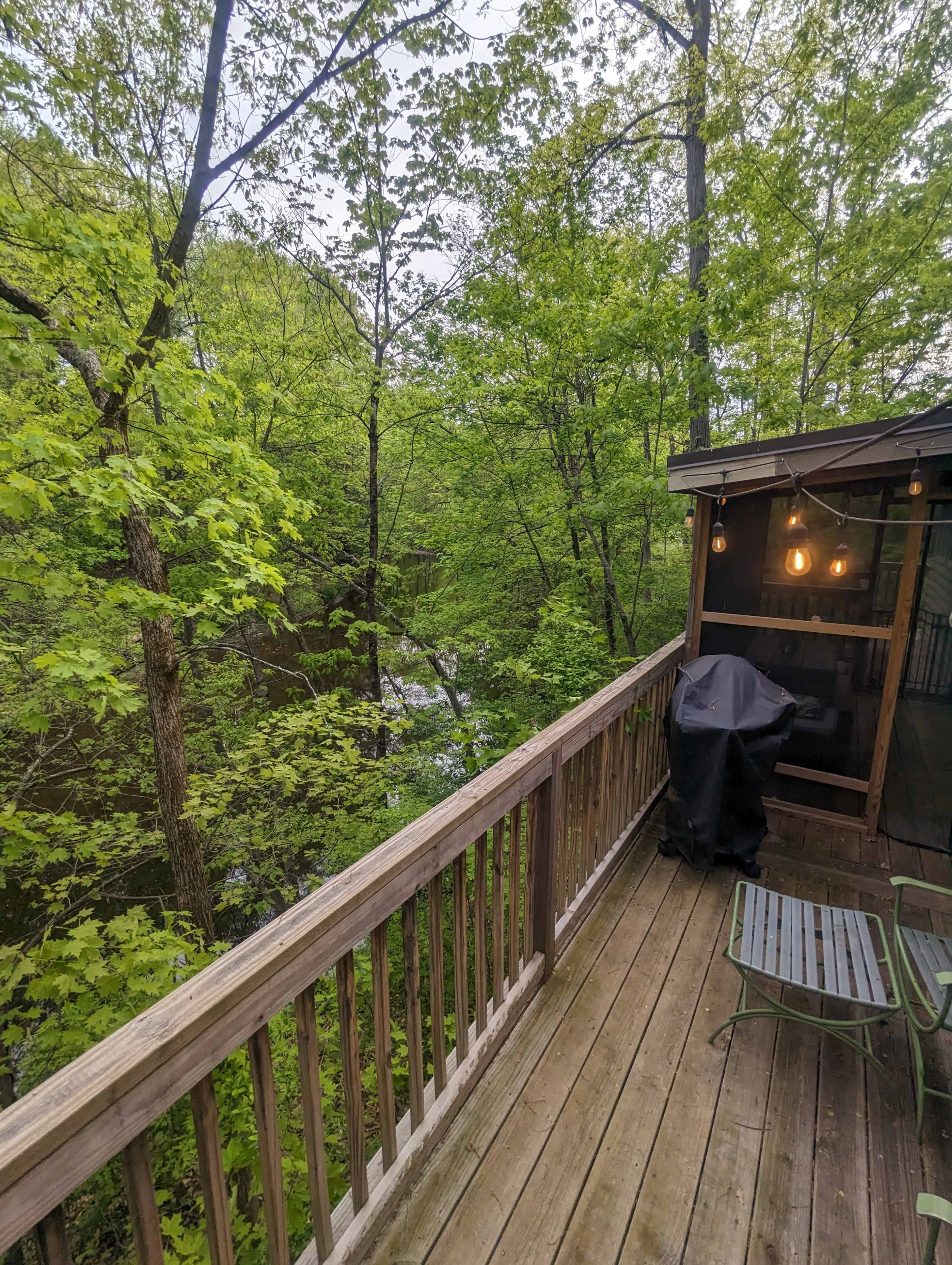 A wooden deck overlooks a leafy green forest and a flowing stream, with a covered grill and a chair positioned beside it.