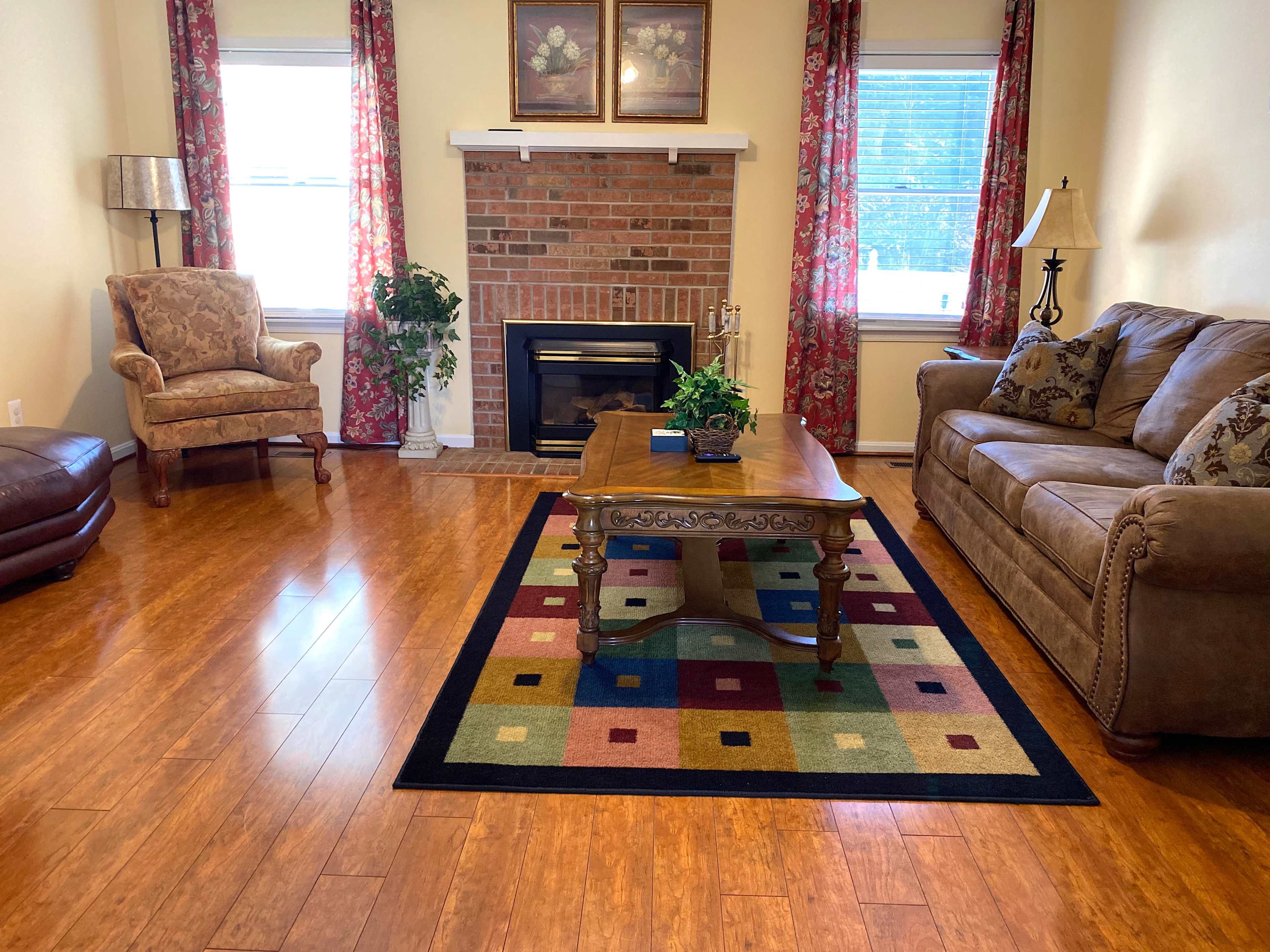 The living room features a brown sofa and armchair, a wooden coffee table, and a fireplace surrounded by decorative plants and framed pictures.