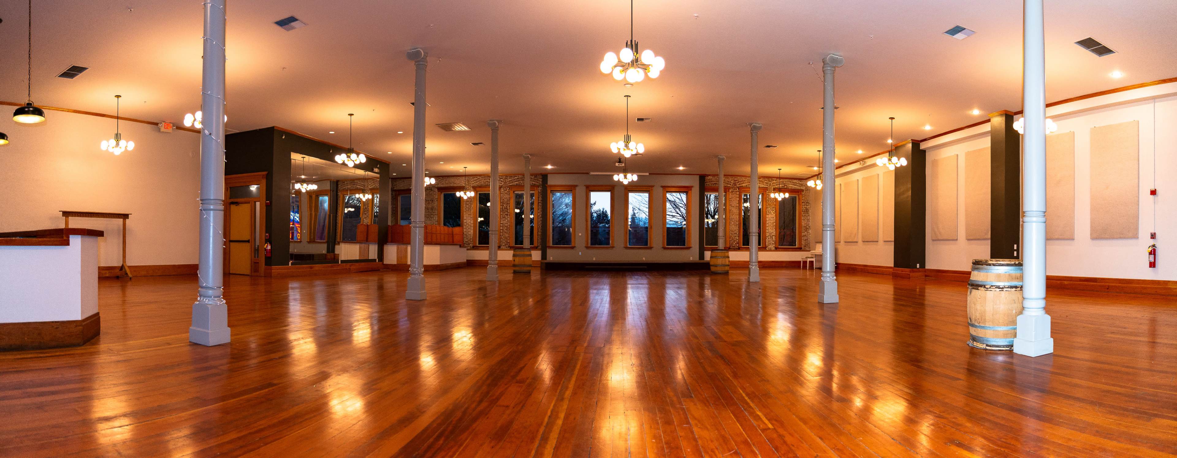 An empty, well-lit event space with wooden flooring, tall ceilings, and large windows.