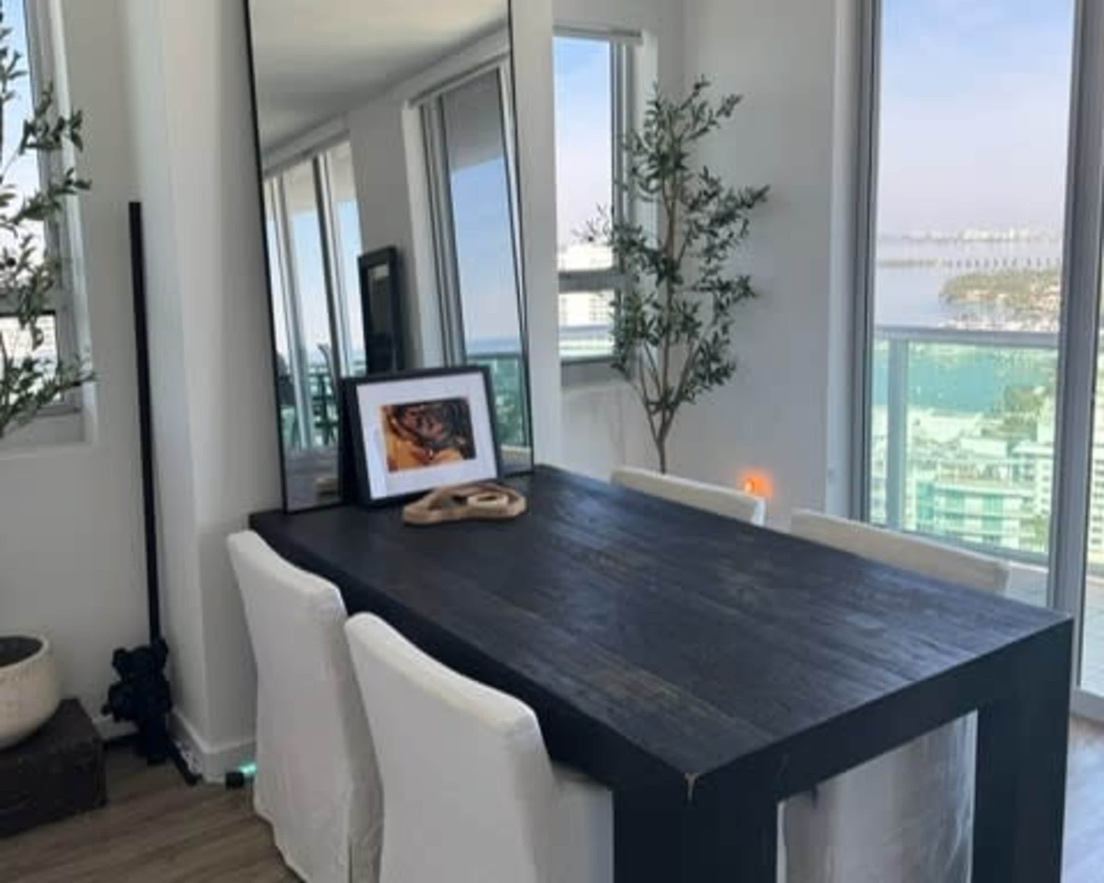 A wooden dining table with white chairs is set against a large mirror and windows, overlooking a cityscape and body of water.