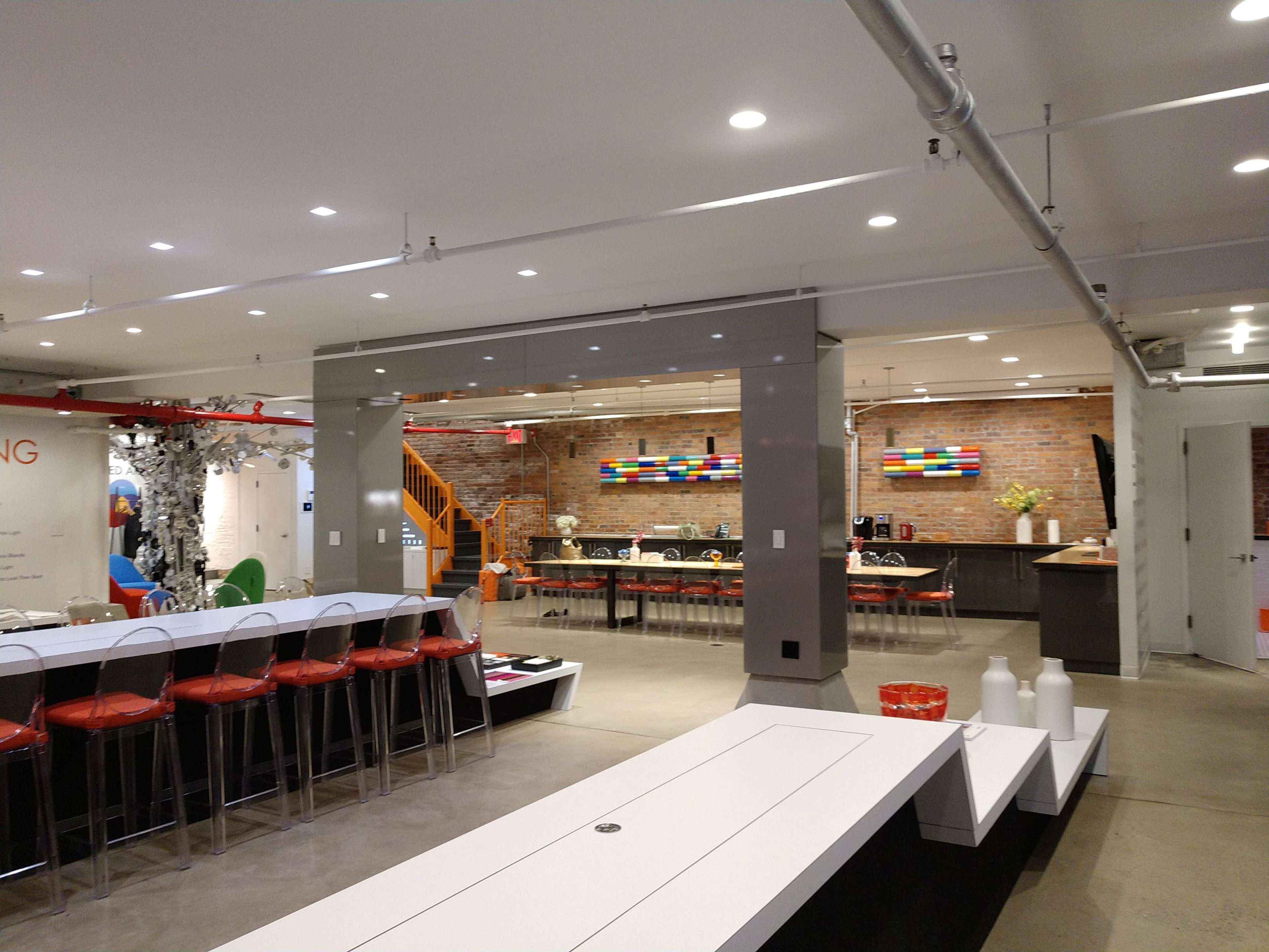 The image shows a modern open-concept office space with a large communal table in the foreground, seating, and a kitchen area in the background featuring brick walls and colorful artwork.