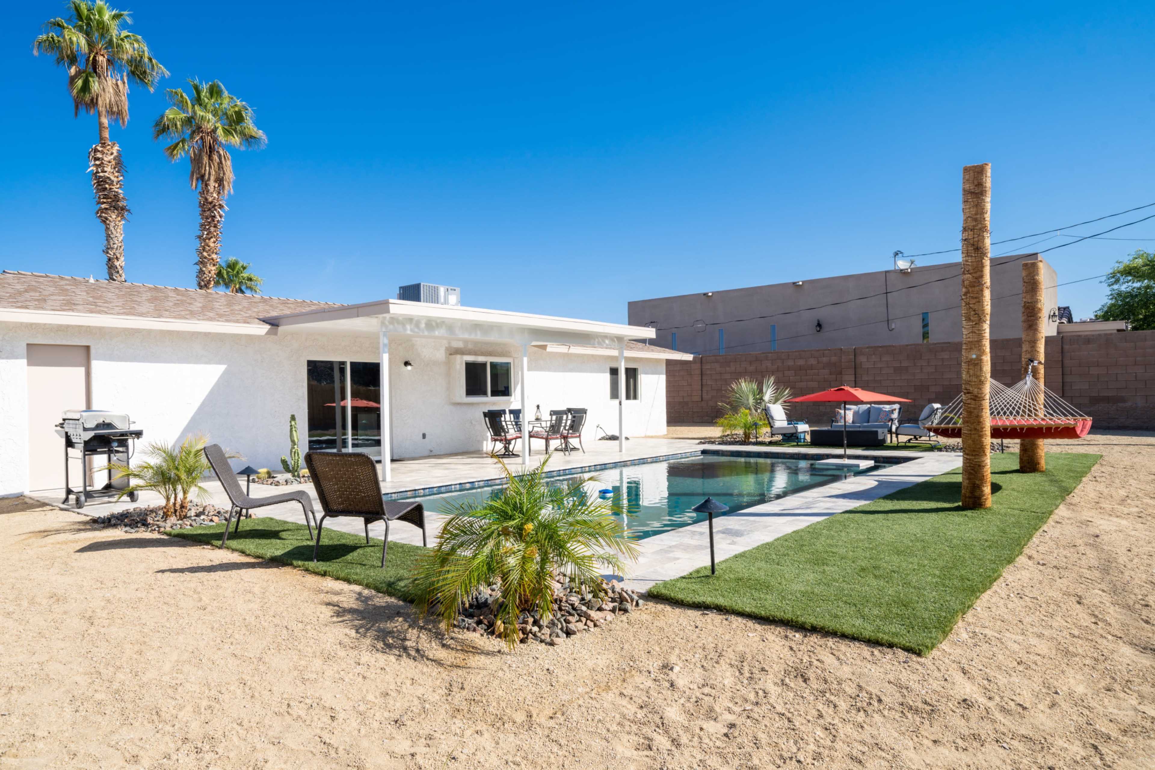 A modern home with a swimming pool is surrounded by palm trees and outdoor furniture in a sandy backyard.