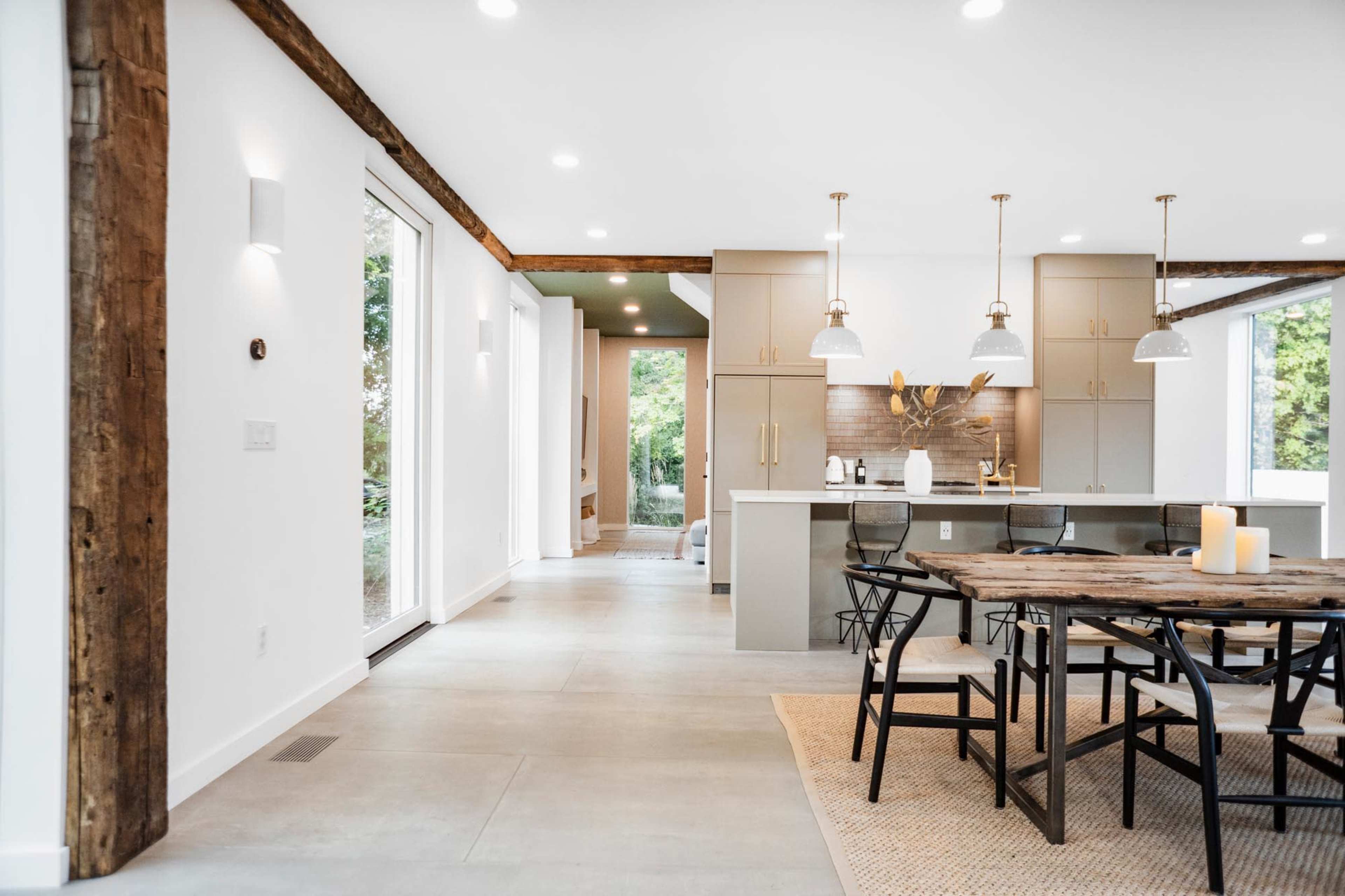 The image shows a modern, open-concept kitchen and dining area with wooden beams, a large wooden table, and minimalist decor.
