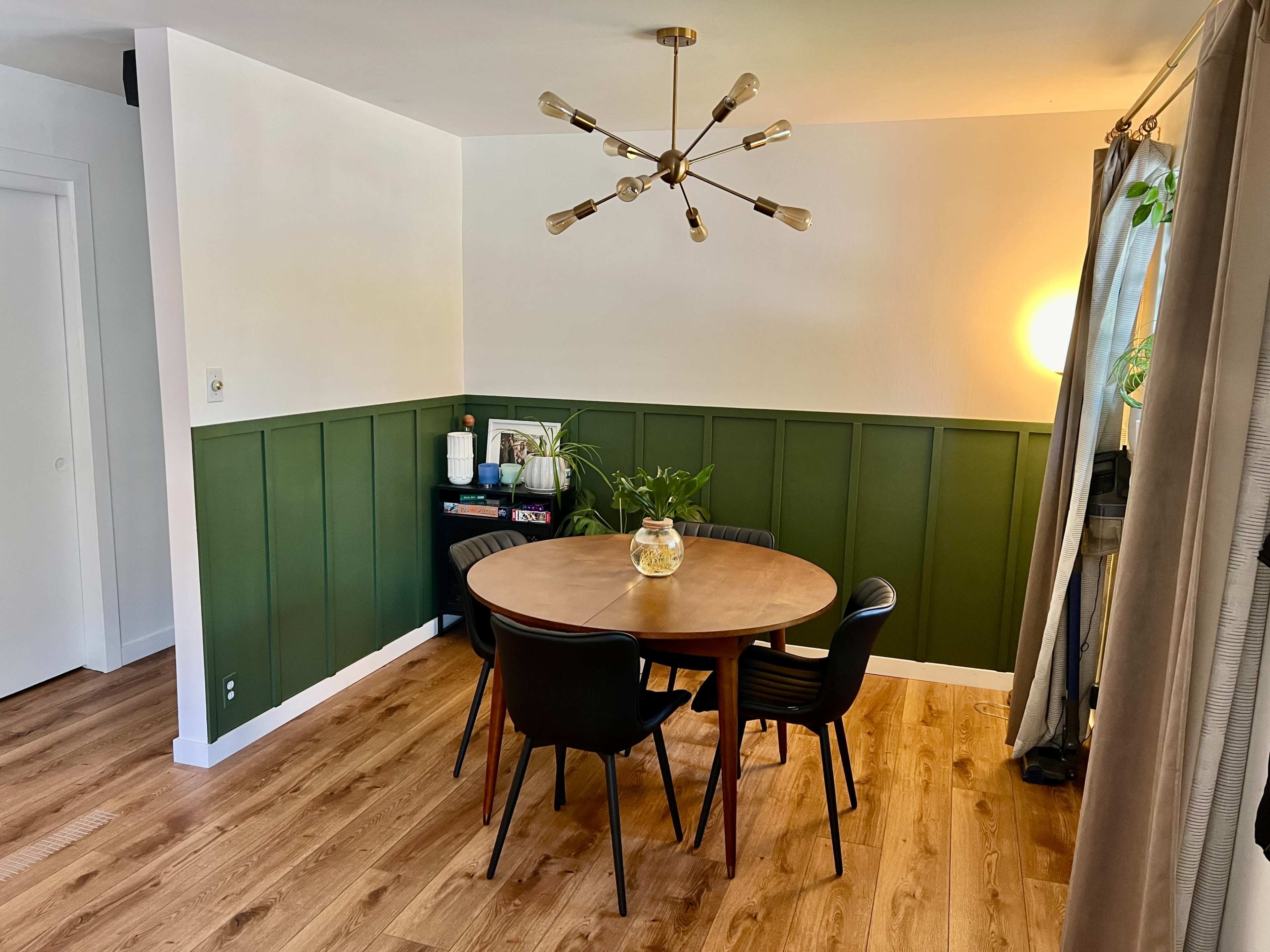 The image shows a round wooden dining table with black chairs surrounded by green-paneled walls and a ceiling light fixture.