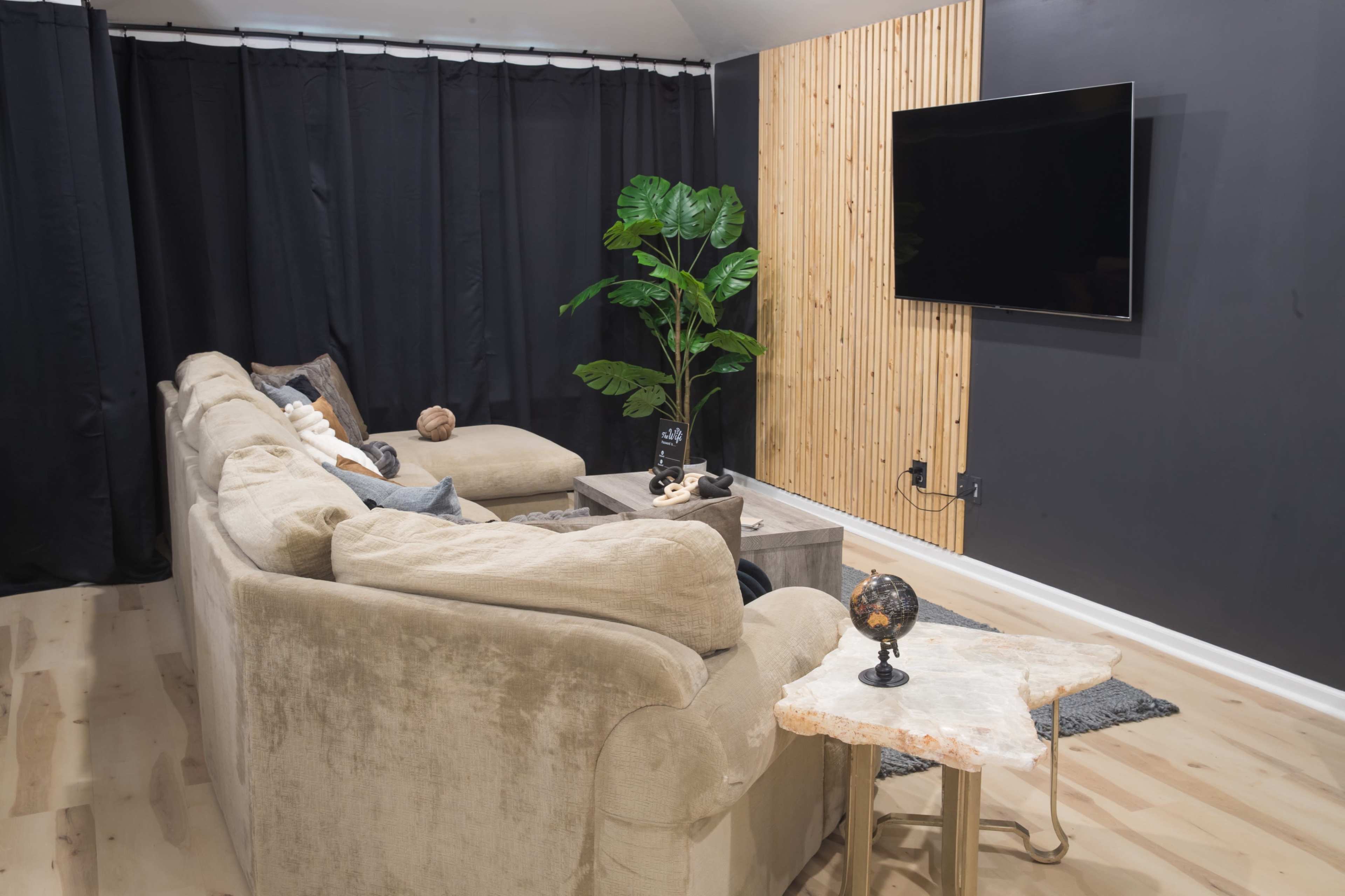 A modern living room with a large, beige sectional sofa facing a wall-mounted television, accompanied by a small side table and a potted plant.