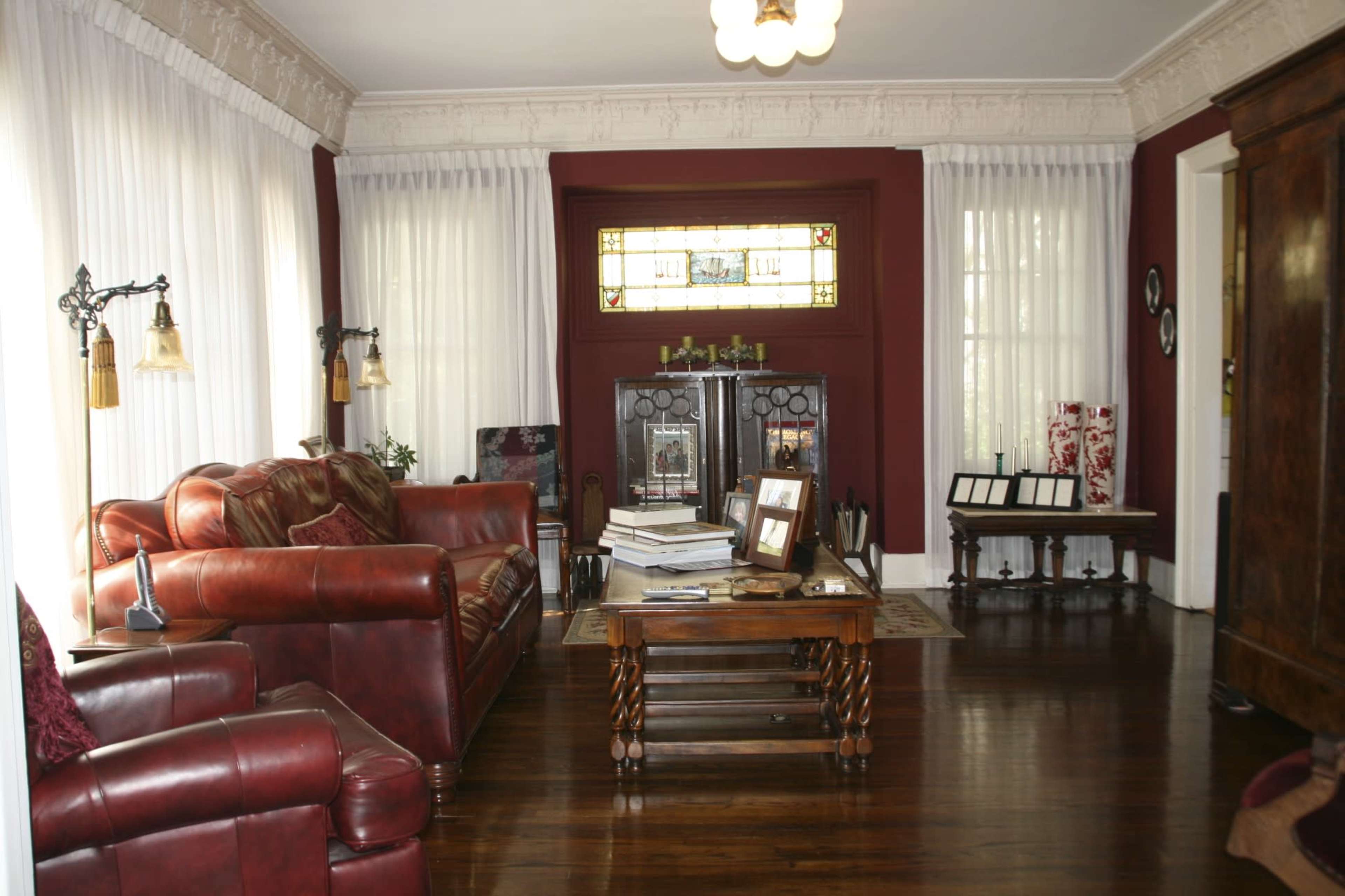 The image depicts a living room with a red leather sofa, a wooden coffee table, large windows dressed with sheer curtains, and decorative cabinets against a dark red wall.