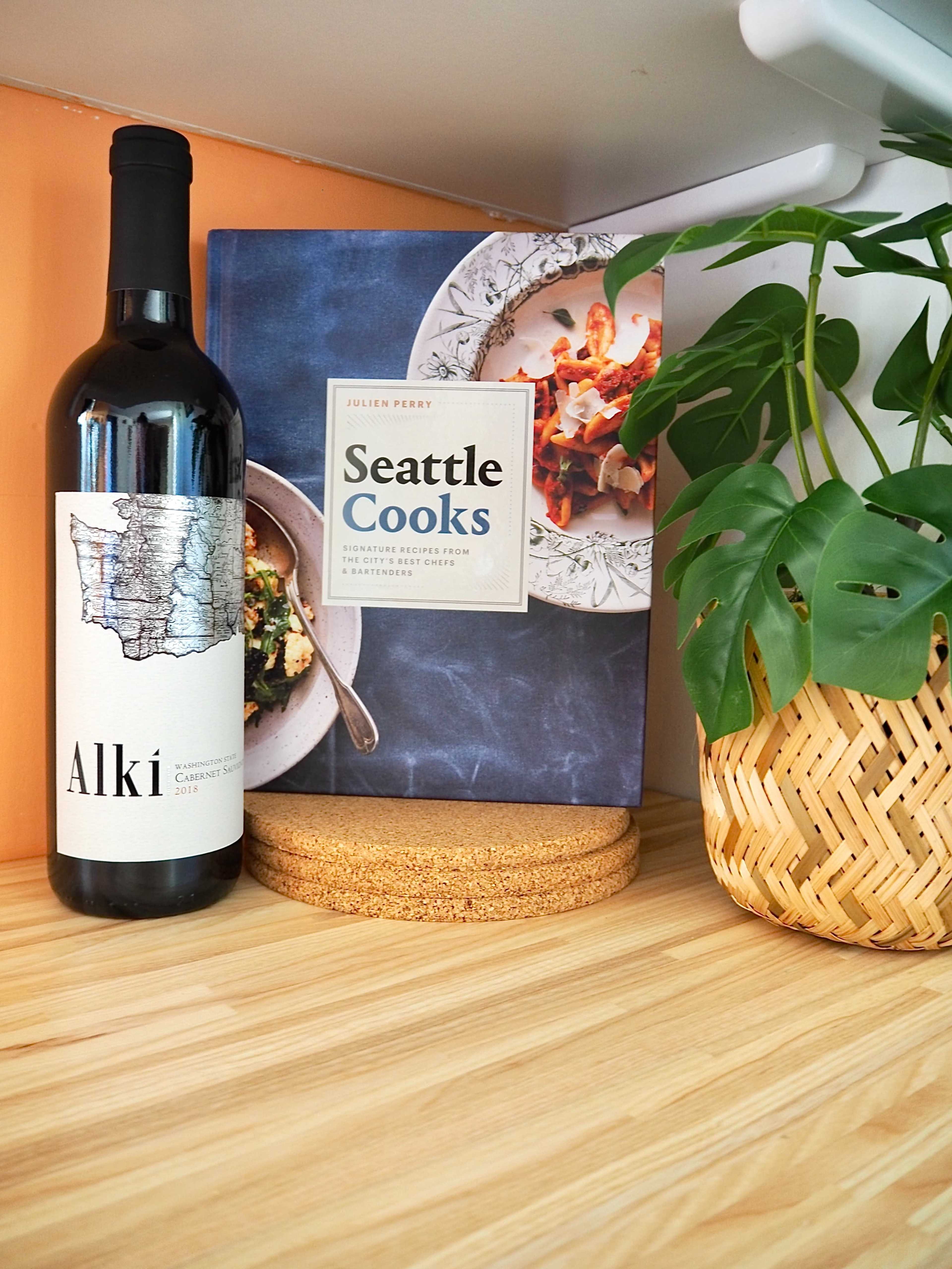 A bottle of red wine labeled "Alki" is placed next to a cookbook titled "Seattle Cooks" on a wooden shelf, accompanied by a woven basket and a Monstera plant.
