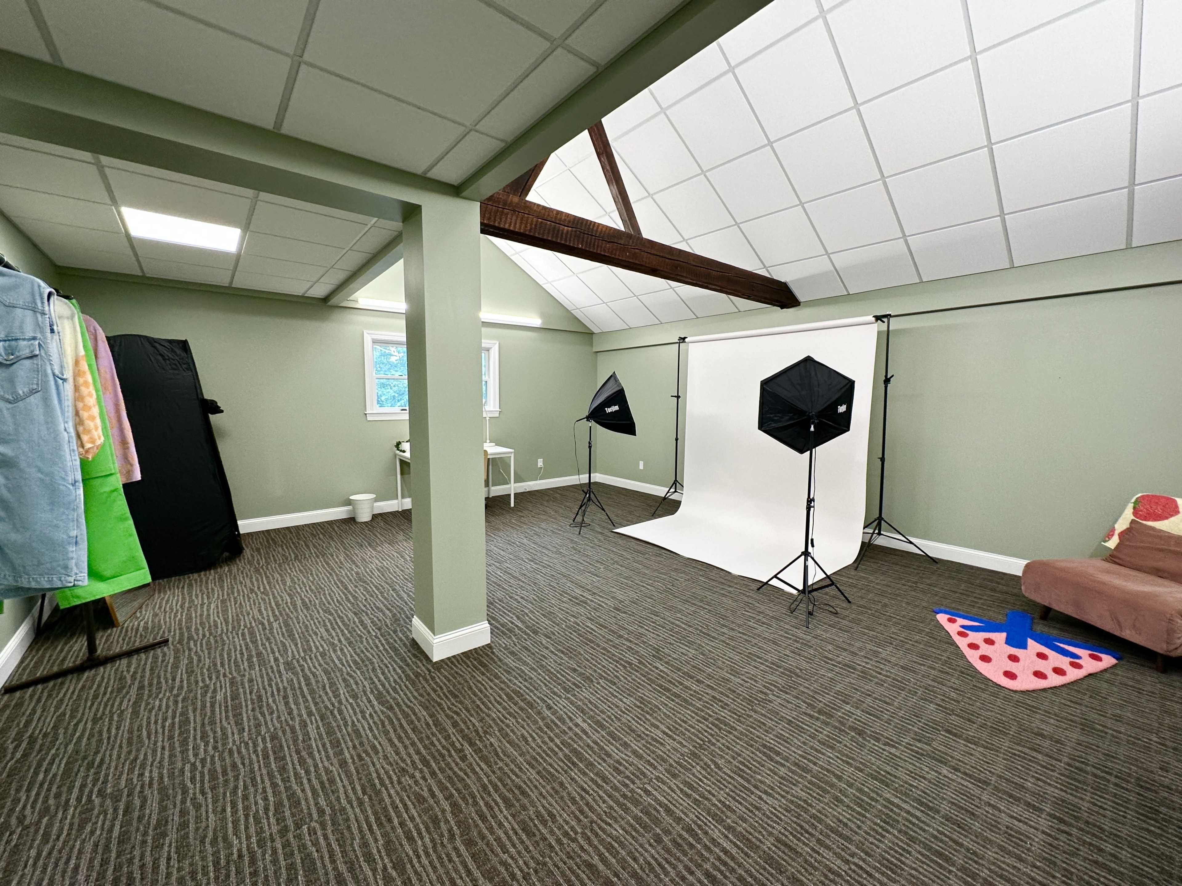 A spacious photo studio with a white backdrop, two softbox lights, a couch, clothing racks, and a colorfully patterned mat on the floor.