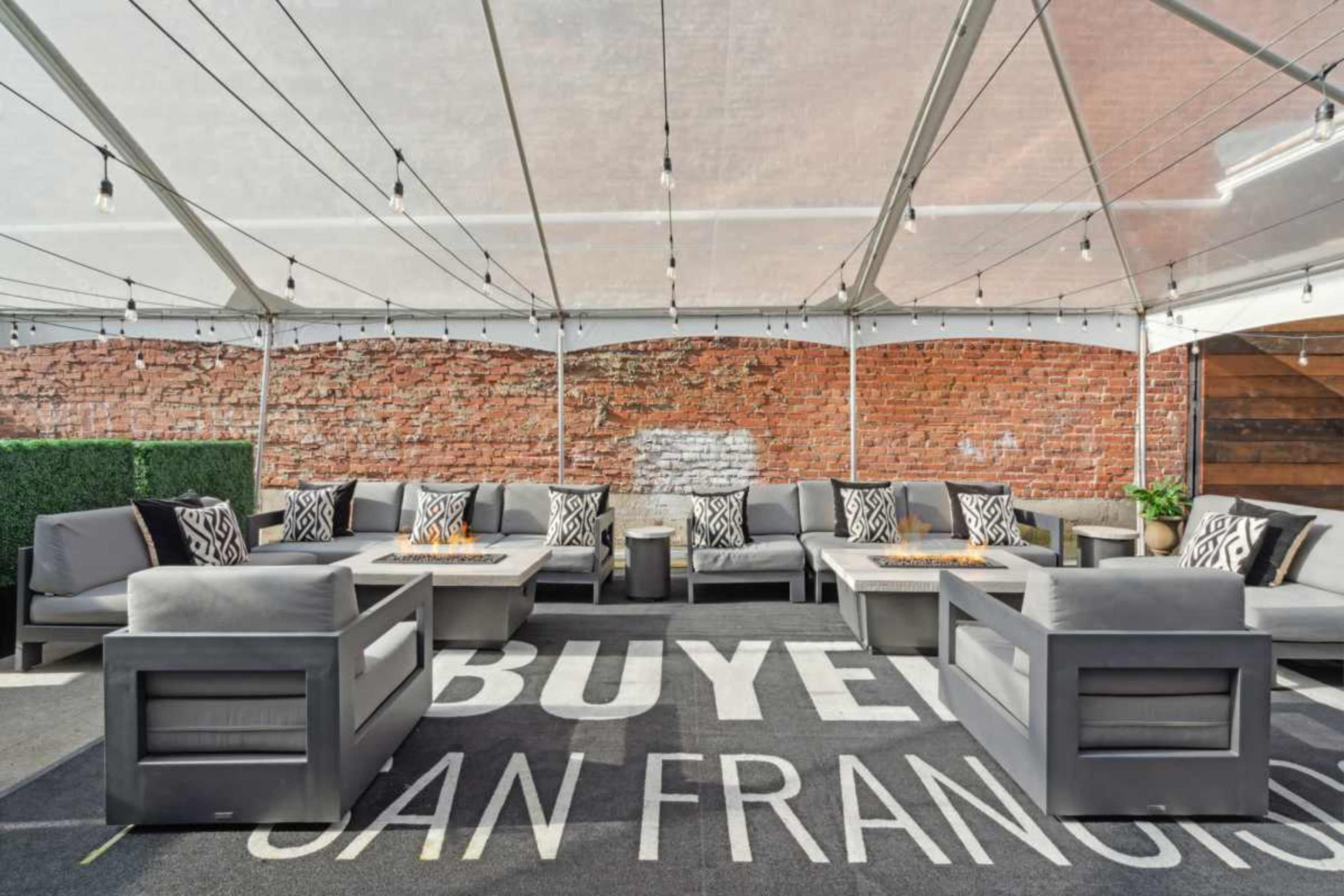 The image shows a tented outdoor lounge area with grey sofas, patterned cushions, and a brick wall backdrop.