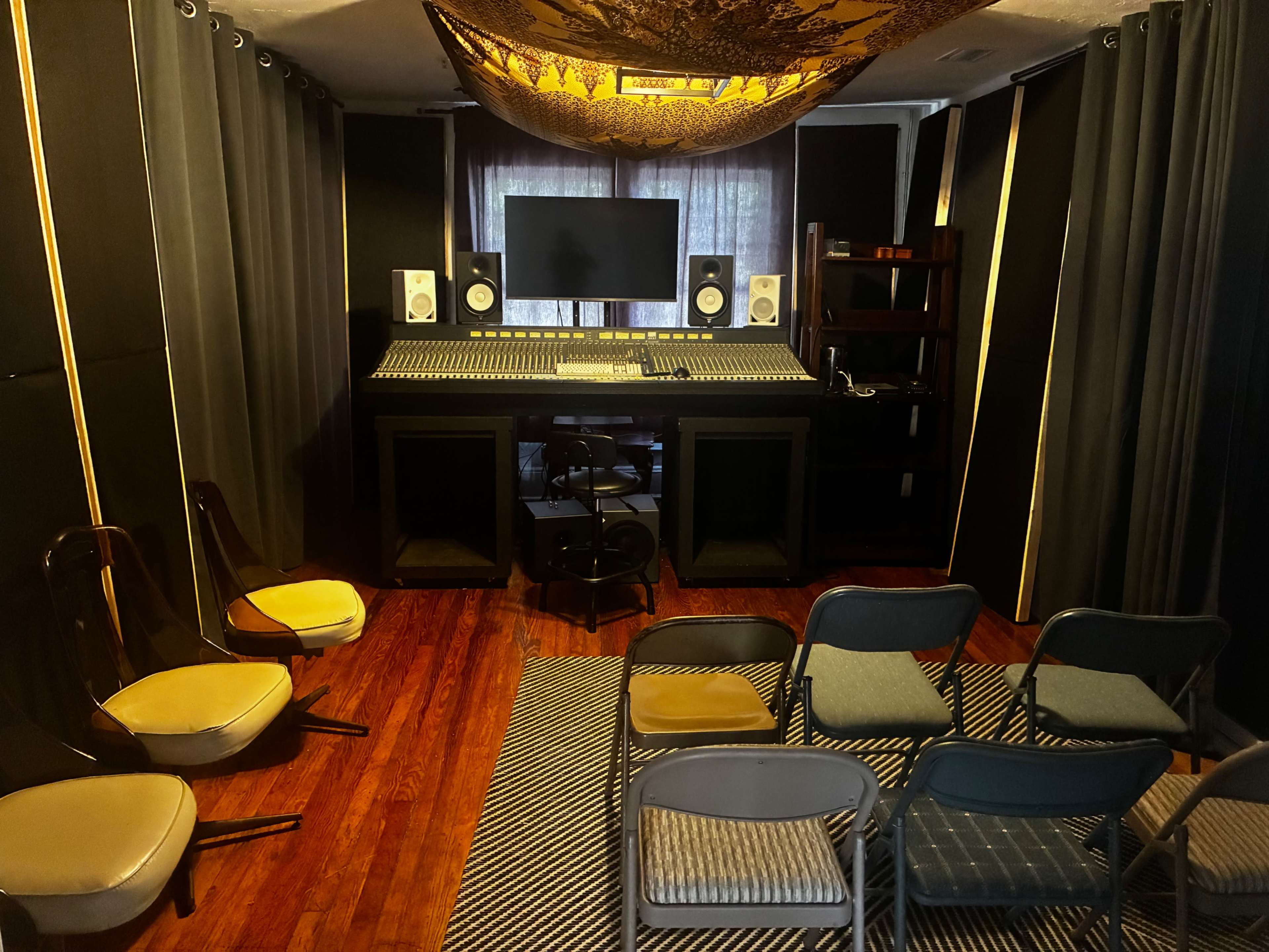 The image shows a sound studio with a mixing console, computer monitor, and several chairs arranged around a striped rug.