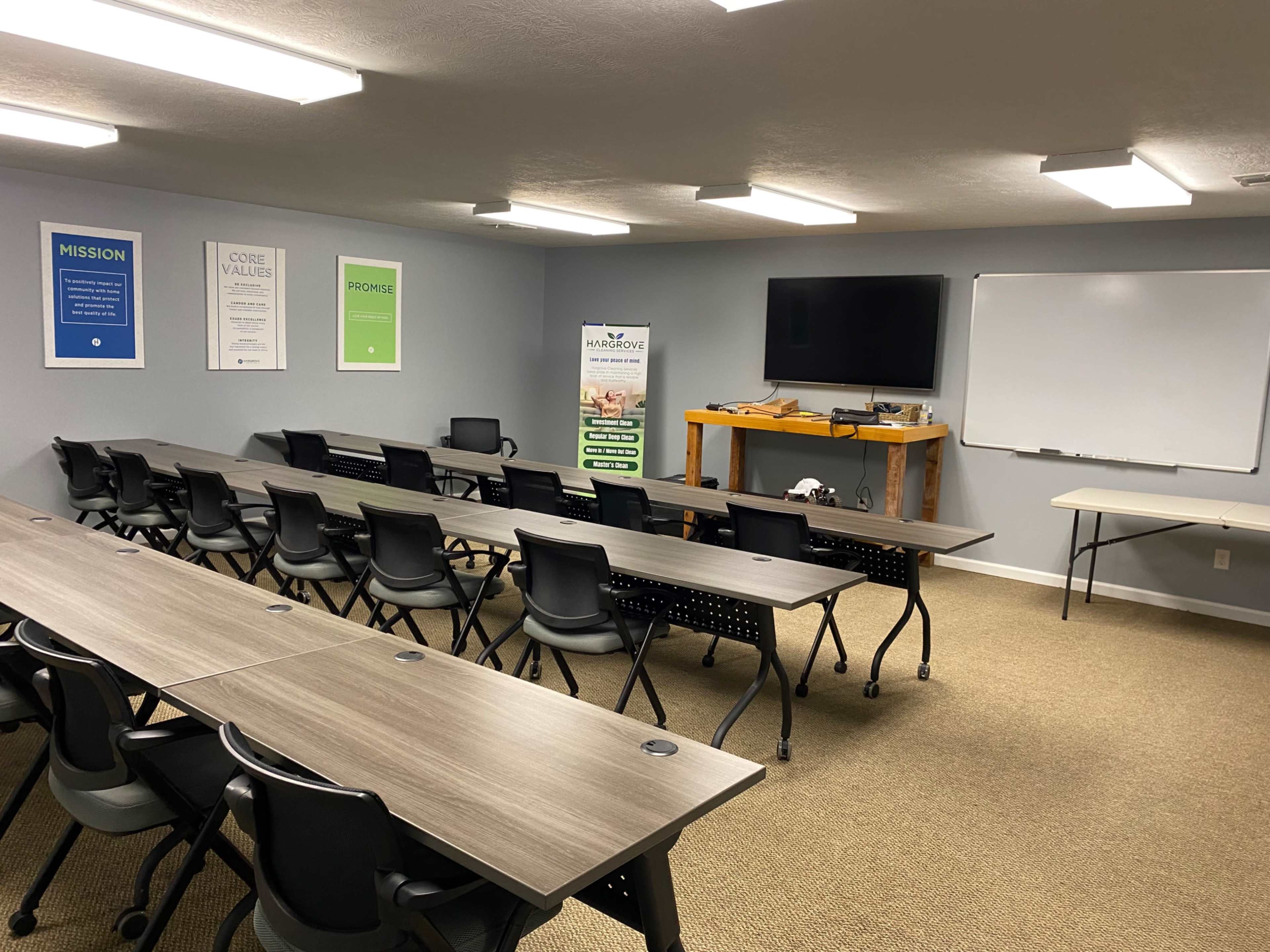 A modern classroom is arranged with multiple rows of tables and chairs, featuring a whiteboard and a television screen at the front, along with motivational posters on the walls.