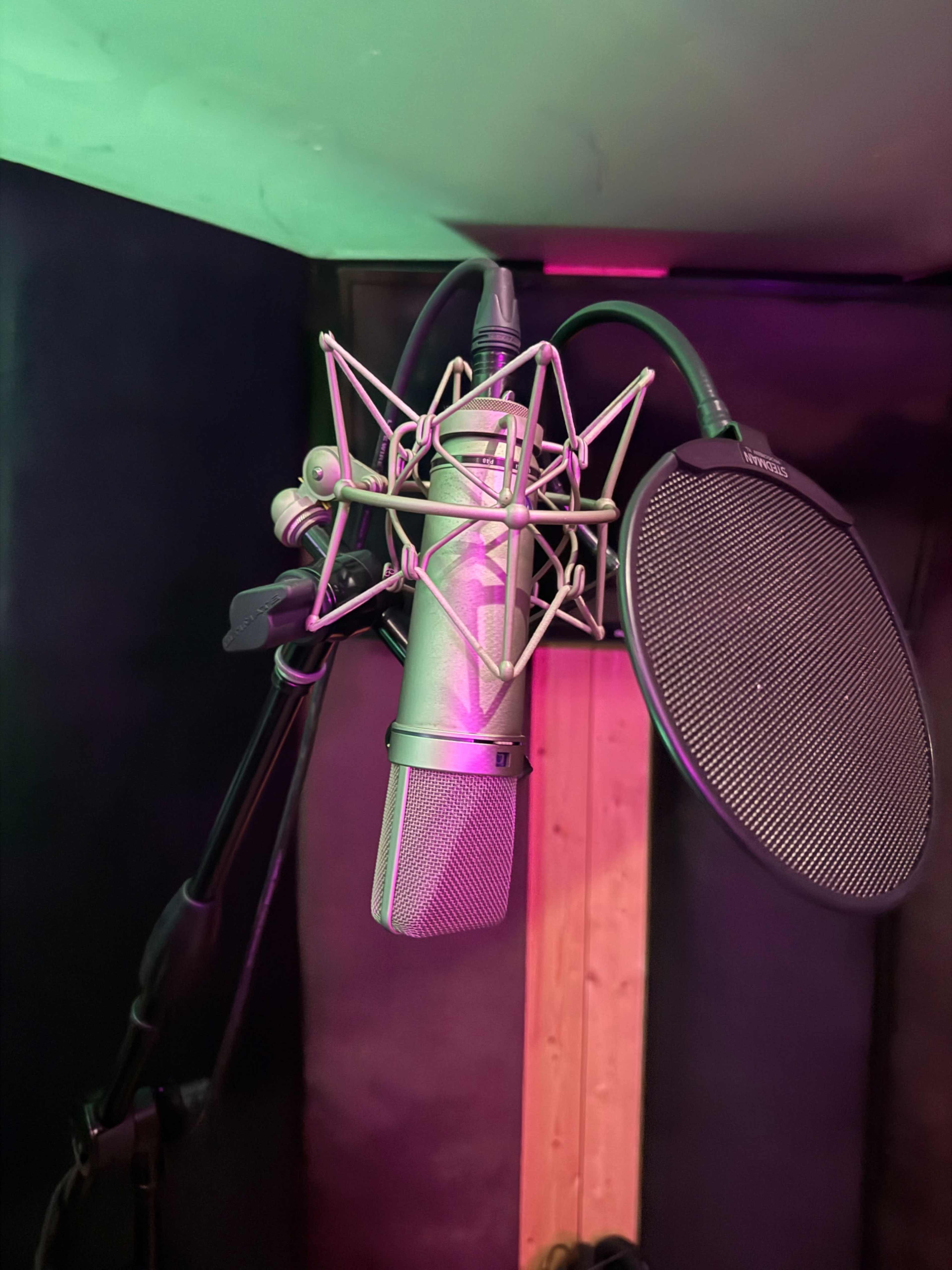 A microphone with a pop filter is mounted in a recording studio corner with dim lighting.