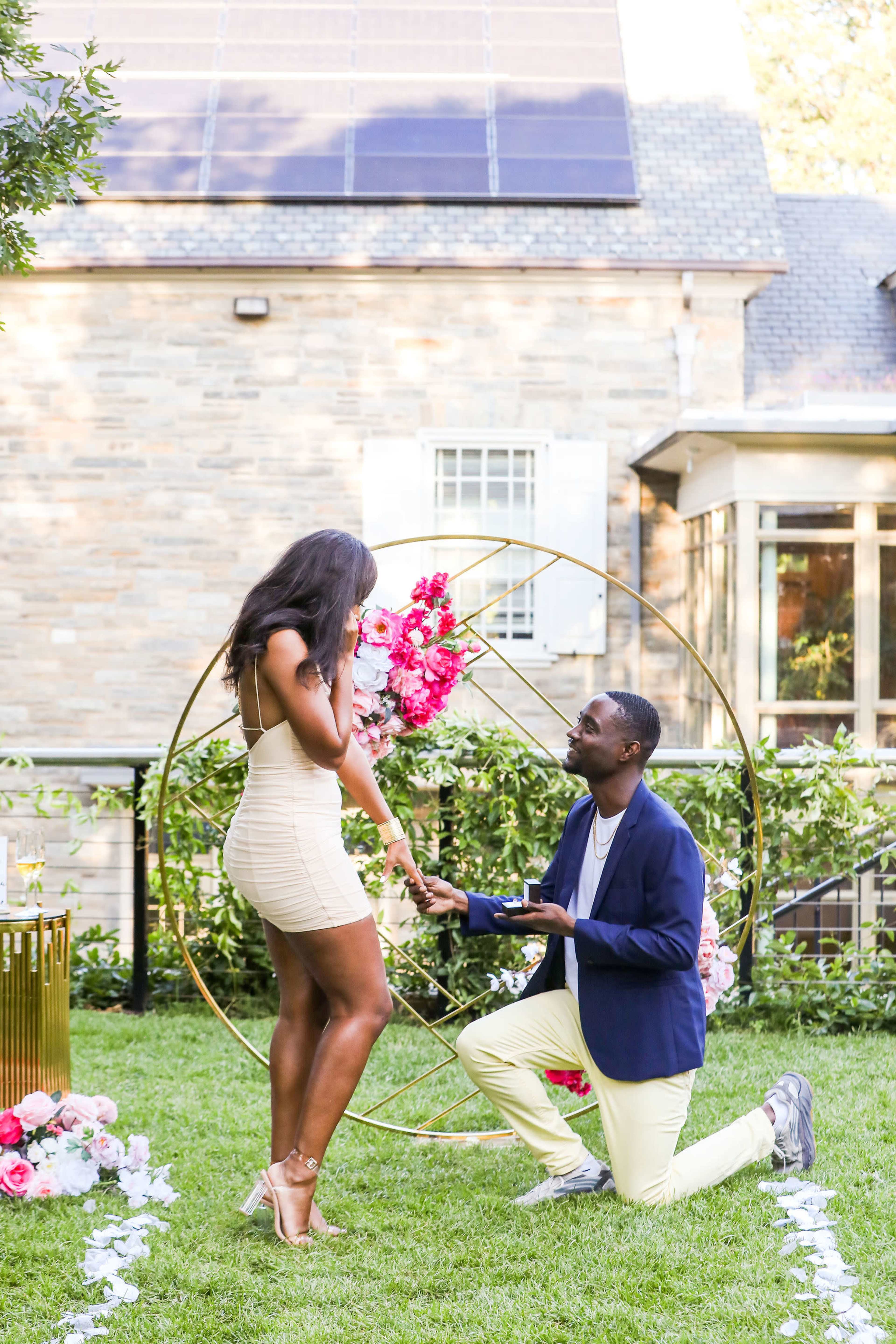 A man kneels on one knee in a garden adorned with flowers, while a woman stands before him, accepting a proposal.