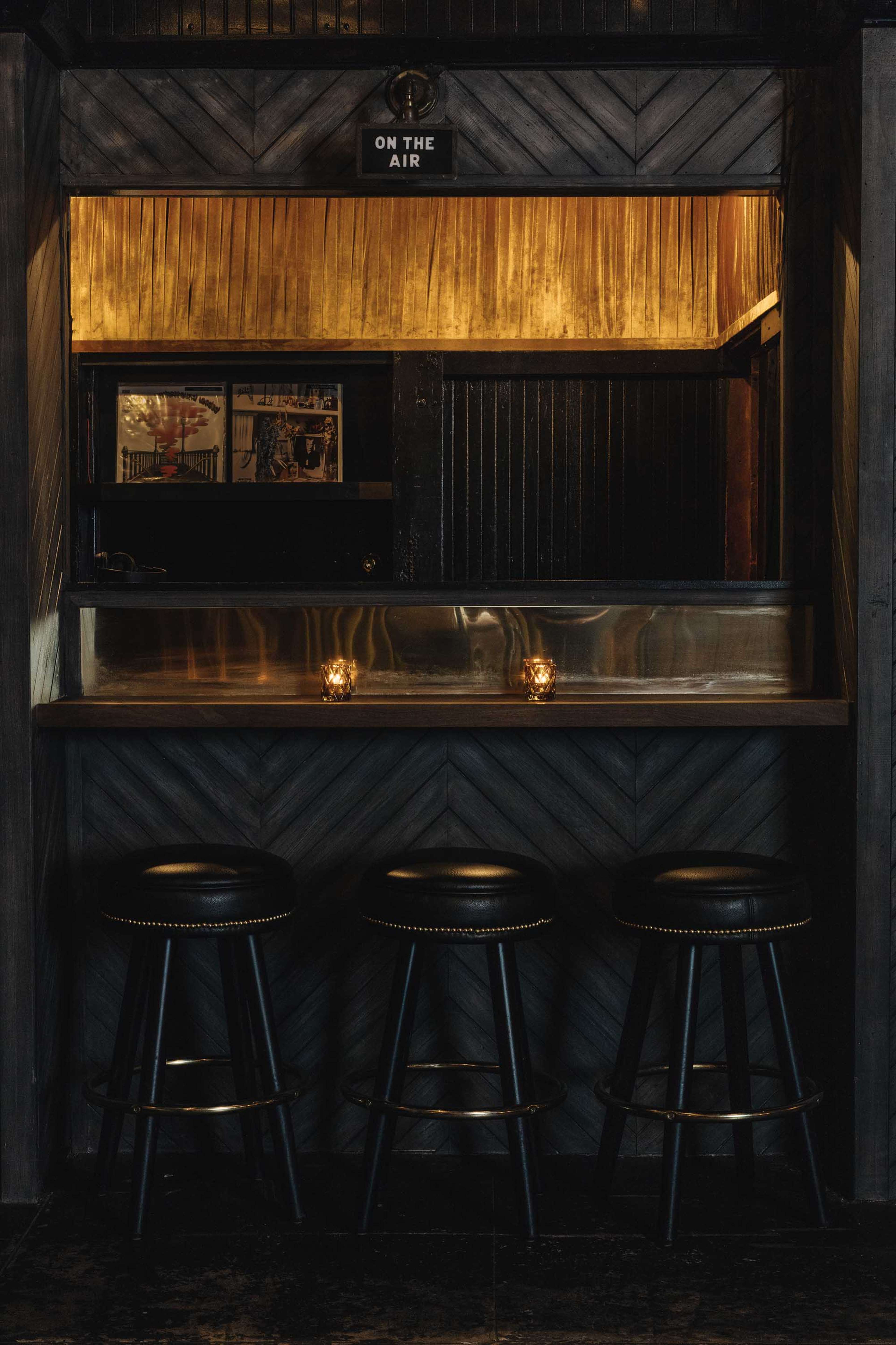 A dimly lit bar area features a wooden counter with three black stools and illuminated candles.