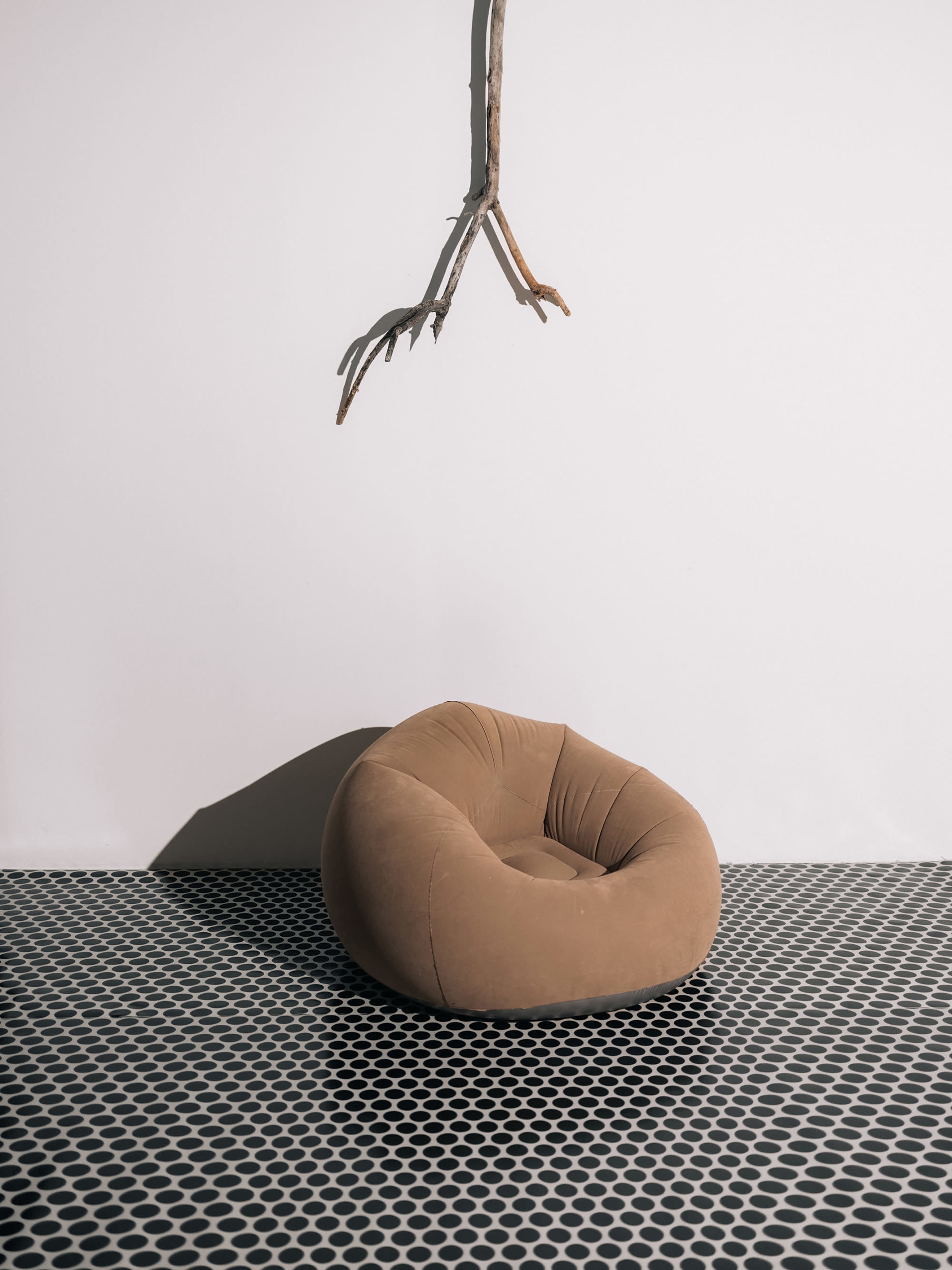 A brown bean bag chair is positioned on a patterned black and white floor beneath a branch hanging from a white wall.