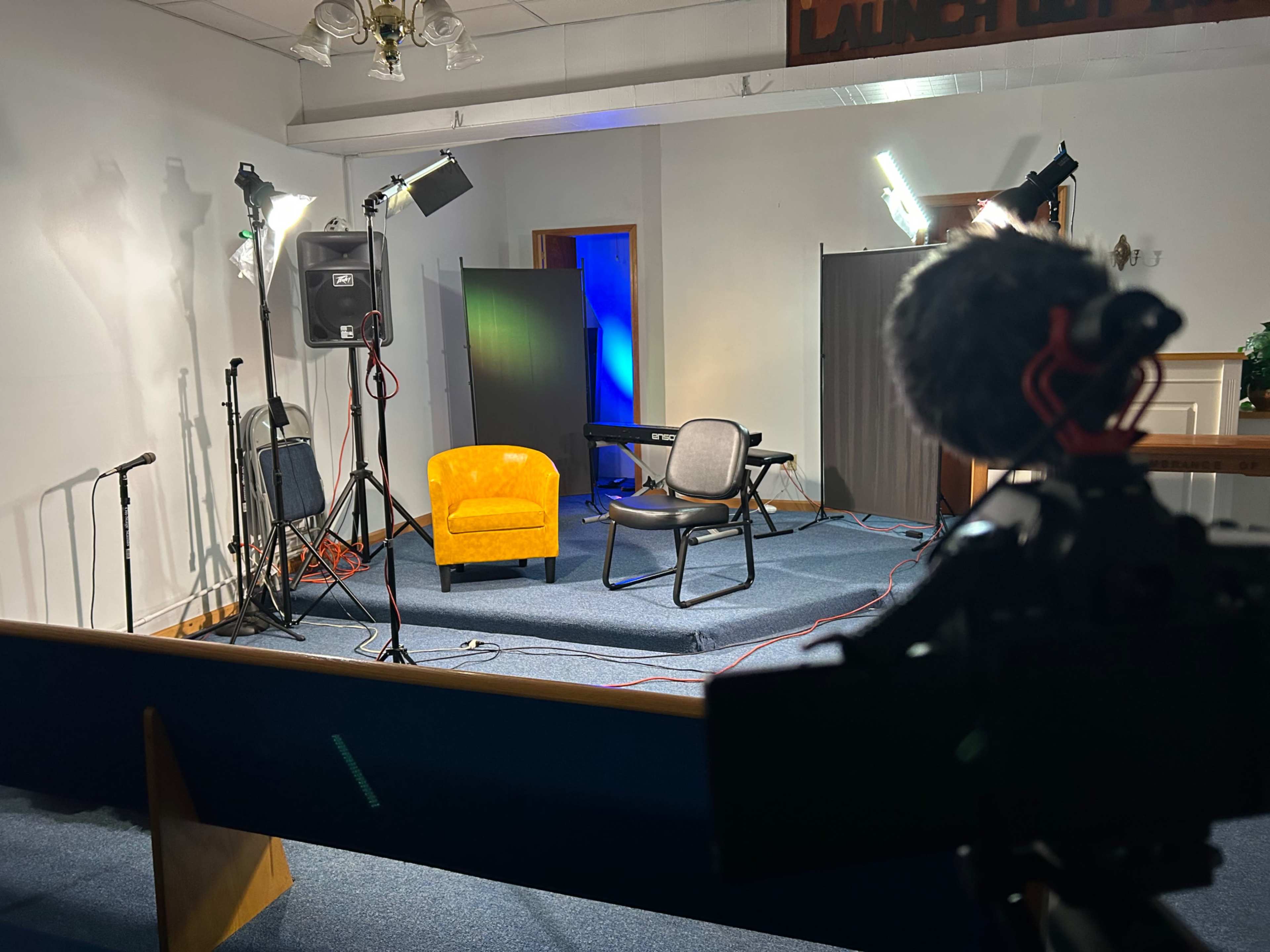A staged interview setup with a yellow armchair, a black chair, microphones, lighting equipment, and a keyboard in a room with a blue backdrop.