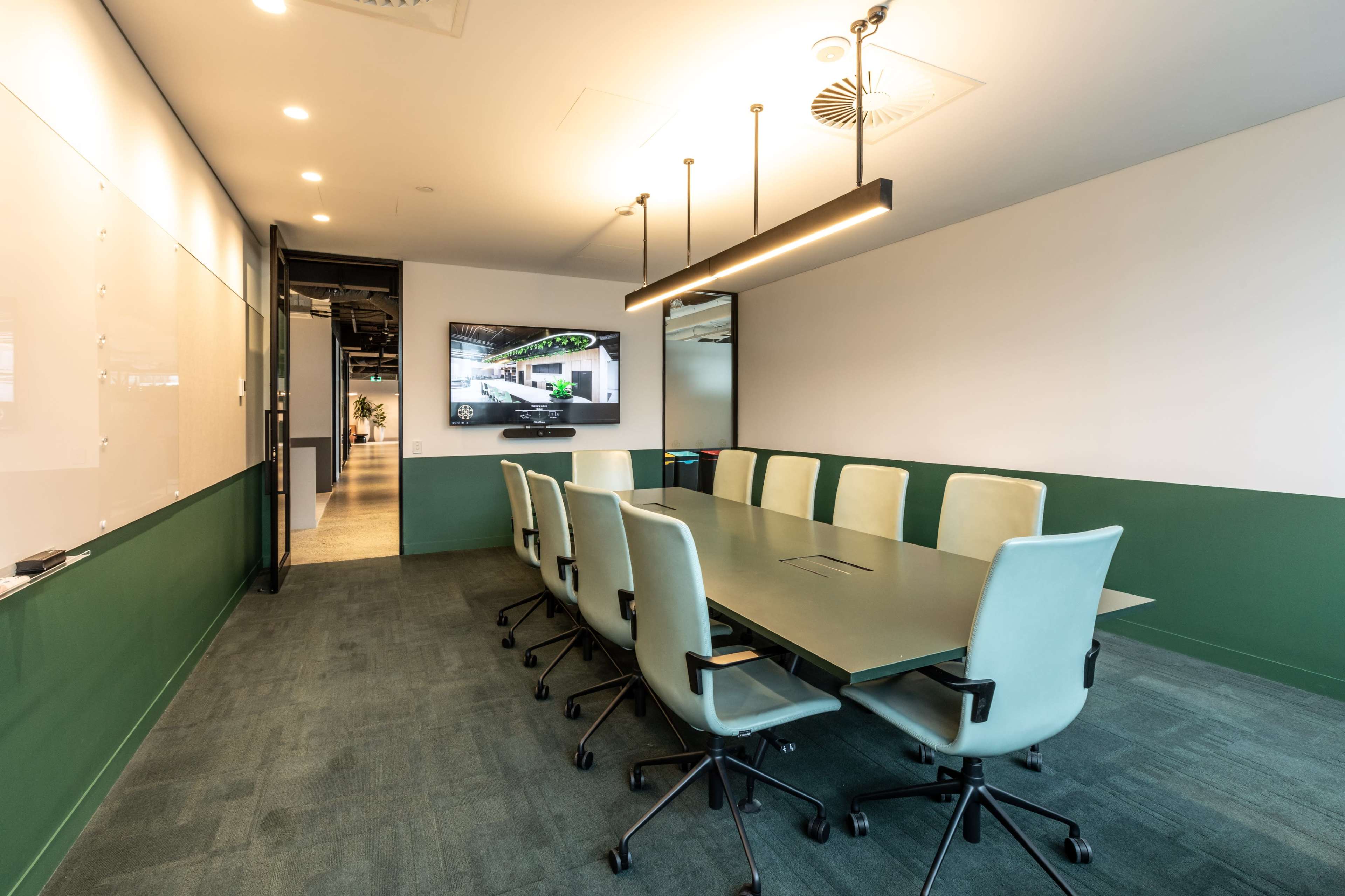 A modern conference room features a long rectangular table surrounded by light green chairs, with a large screen on one wall and a whiteboard on another.