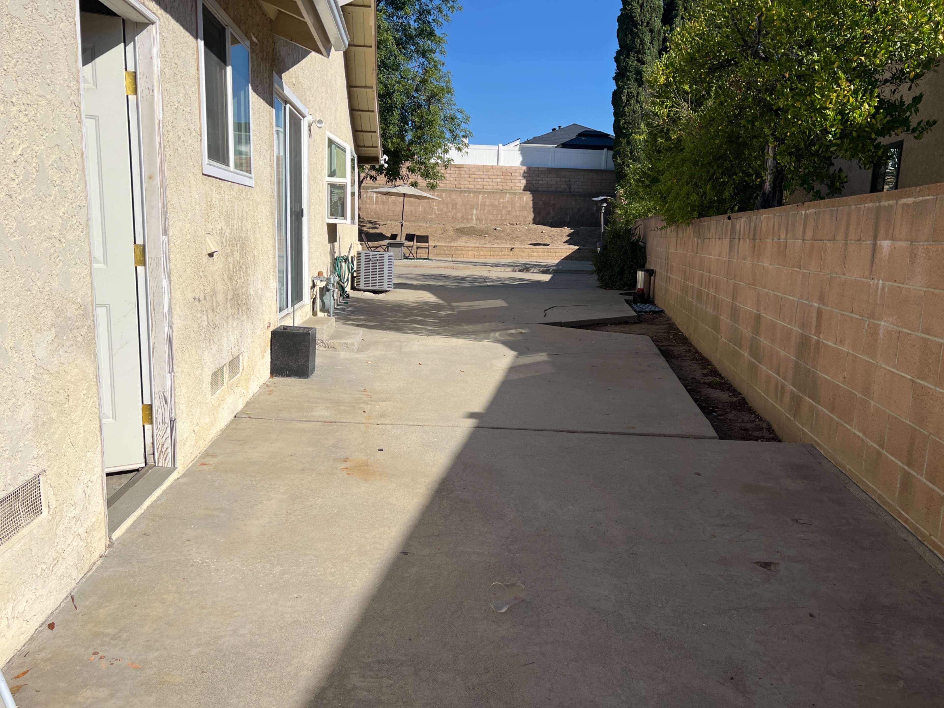 A concrete area alongside a house, bordered by a wall and a row of trees, with a vacant space leading to a stepped landscape in the background.