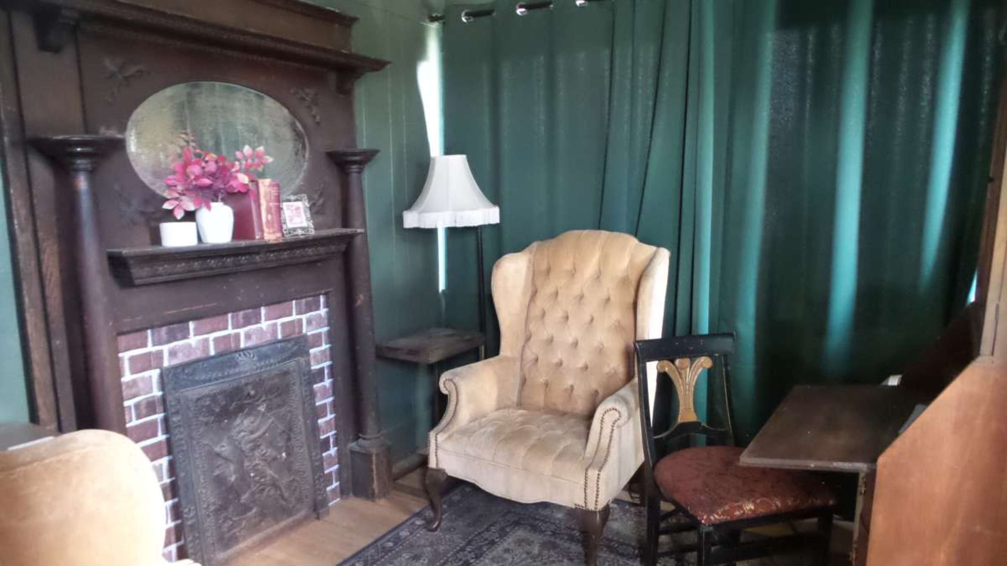 A vintage sitting area with a tufted armchair, a small wooden table, and a fireplace surrounded by a green curtain.