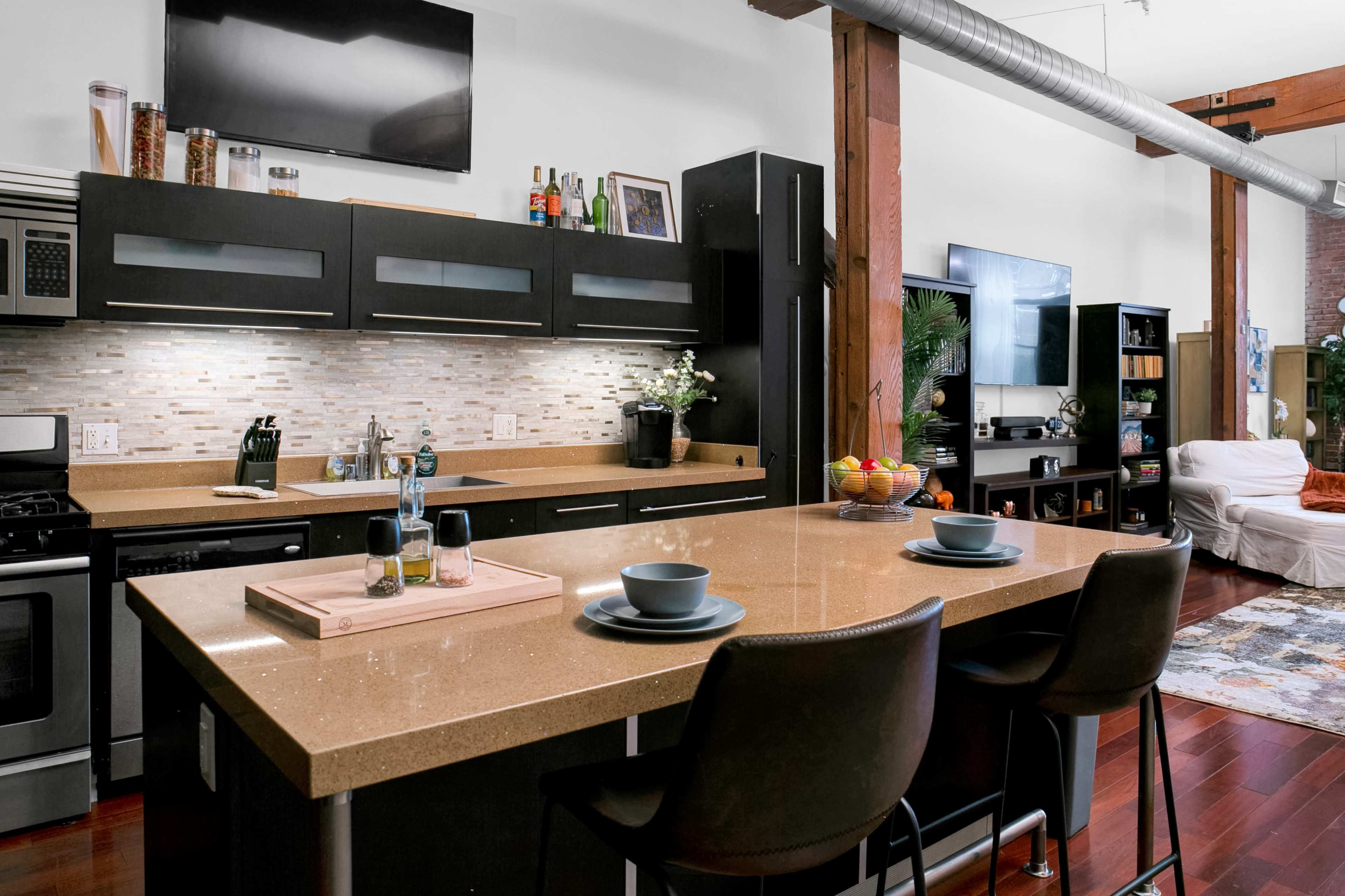 The image shows a modern kitchen with a central island, equipped with two stools, a stovetop, and a variety of kitchen appliances, against a backdrop of exposed wooden beams.