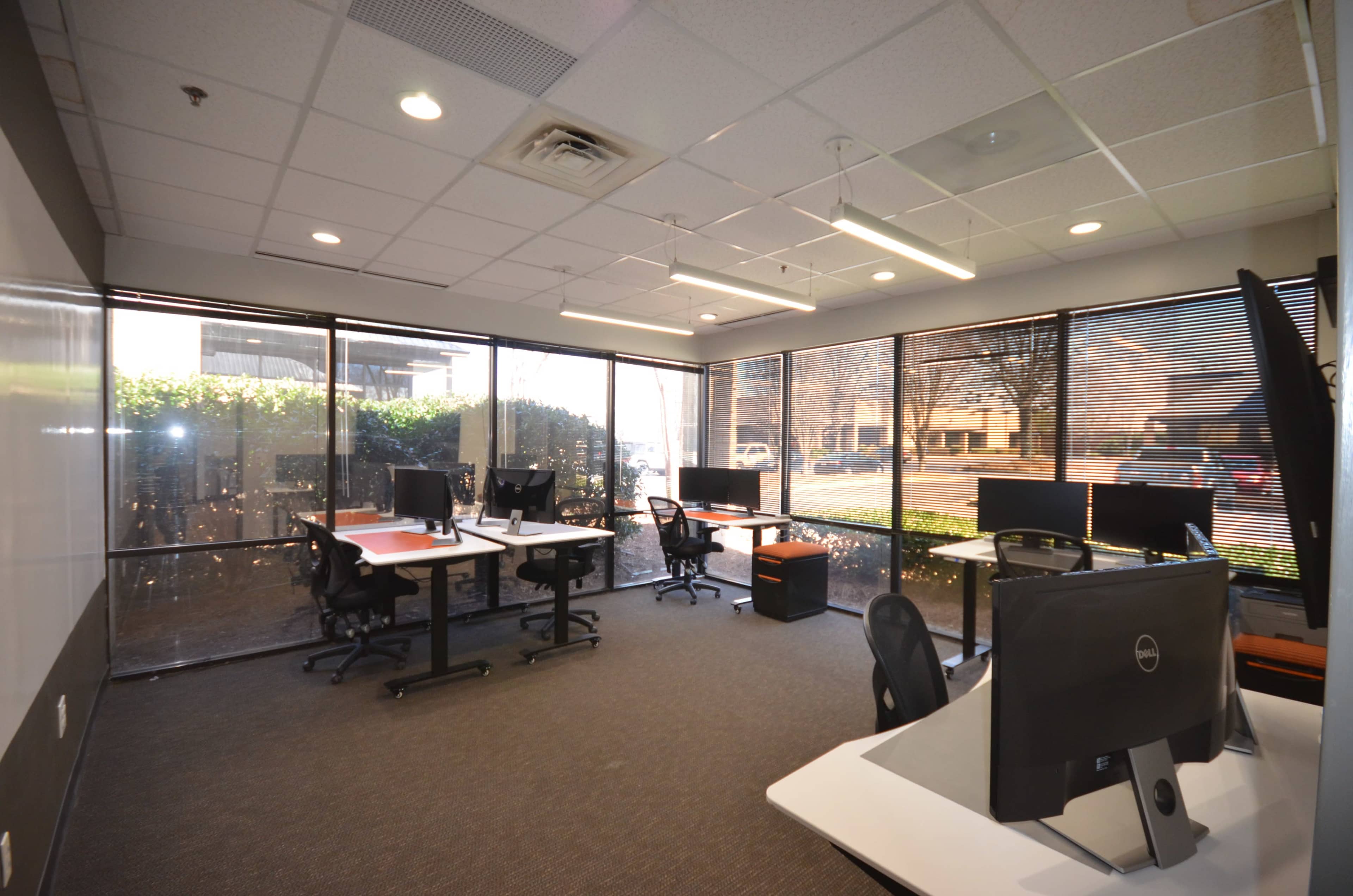 An office space with multiple desks and computer monitors, featuring large windows that provide natural light and a view of the outdoors.