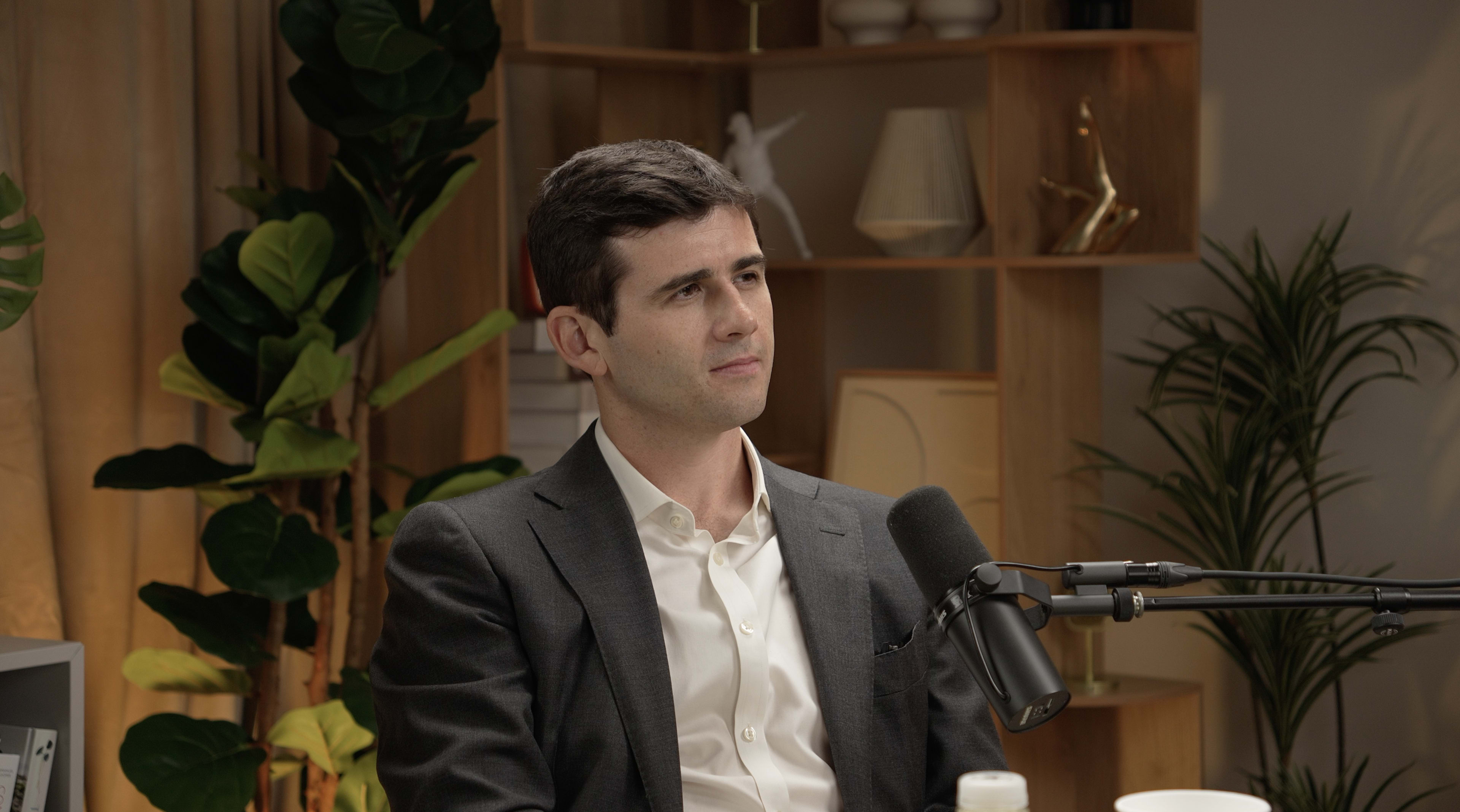 A man in a dark suit sits at a table in a well-lit room with plants and shelves in the background.