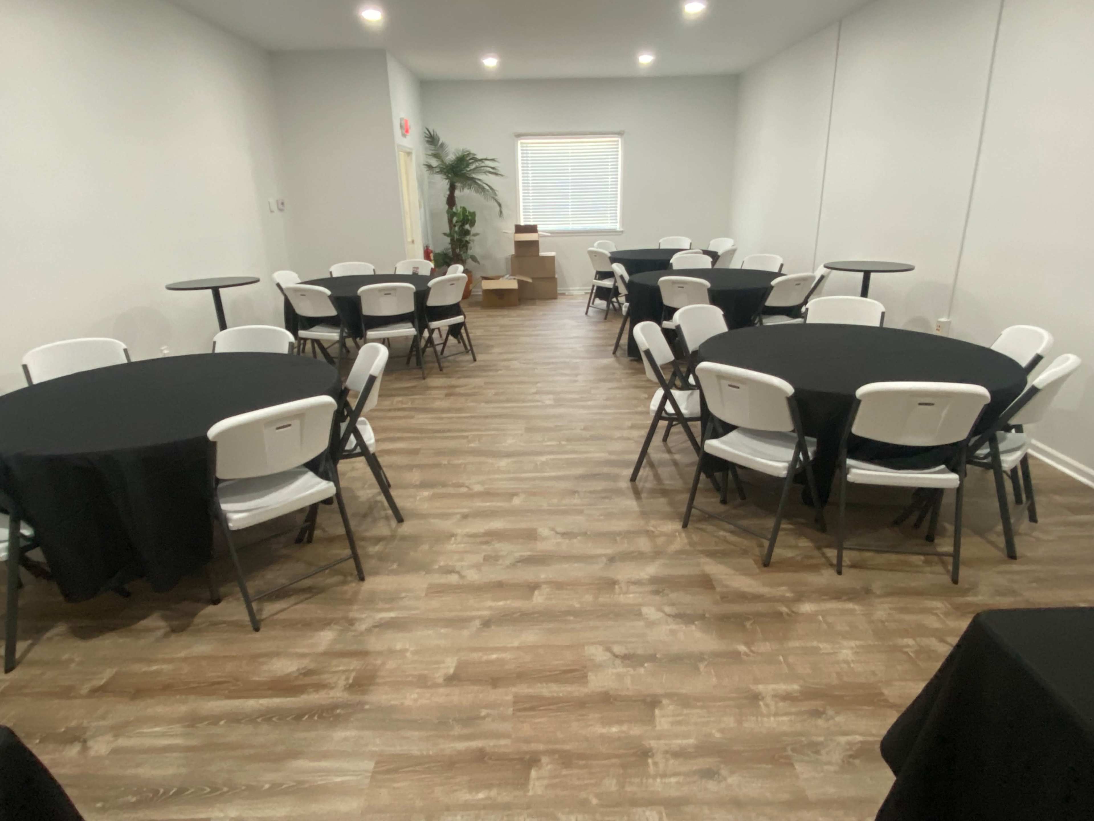 The room features several round tables covered with black tablecloths and white folding chairs, arranged on a wood-like floor with bright lighting and a plant in the corner.